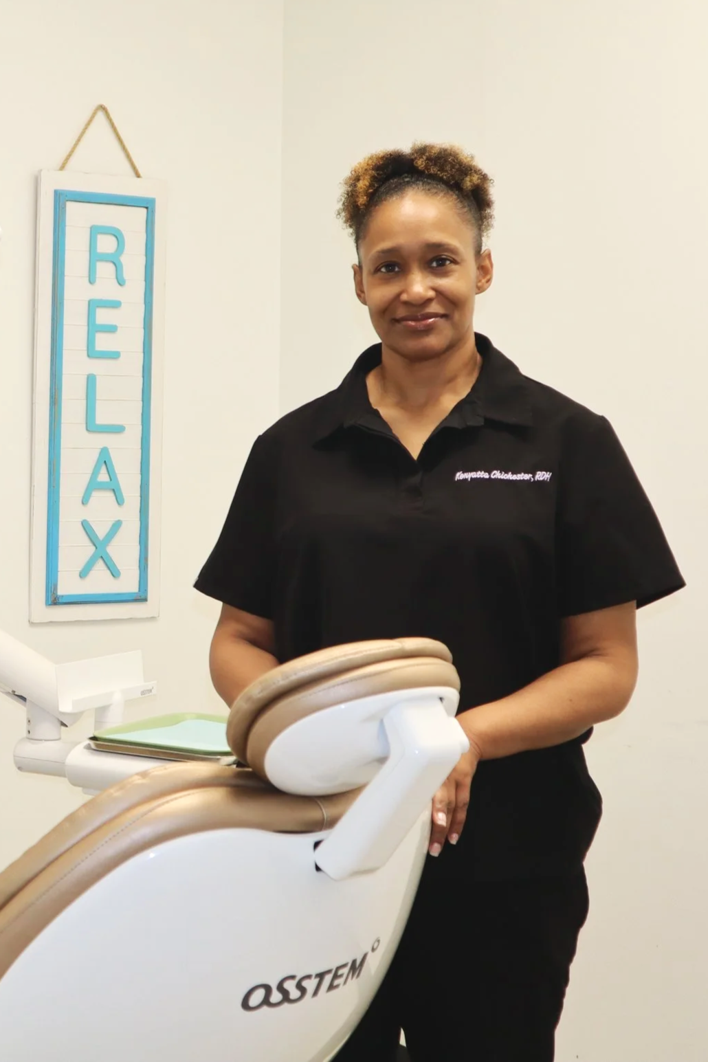 A woman standing next to a dental chair in a clinic, with a blue 'RELAX' sign hanging on the wall behind her.
