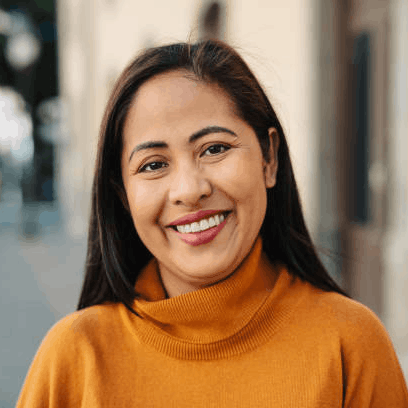 A woman with long dark hair smiling, wearing a mustard yellow turtleneck sweater, standing outdoors on a city street.