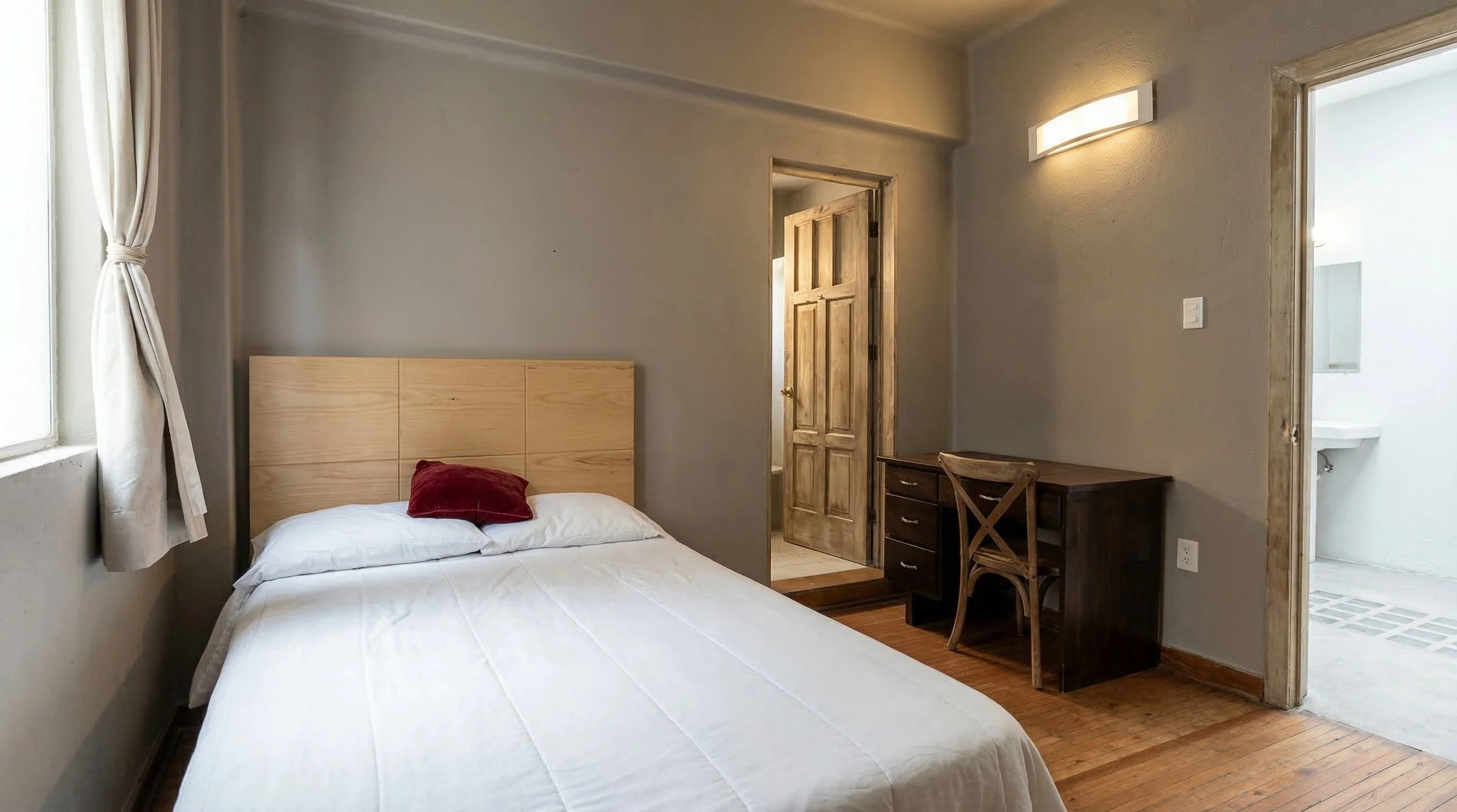 Habitación sencilla con cama de madera, cortinas blancas, escritorio de madera y puerta de madera, con baño visible en la puerta al lado derecho.