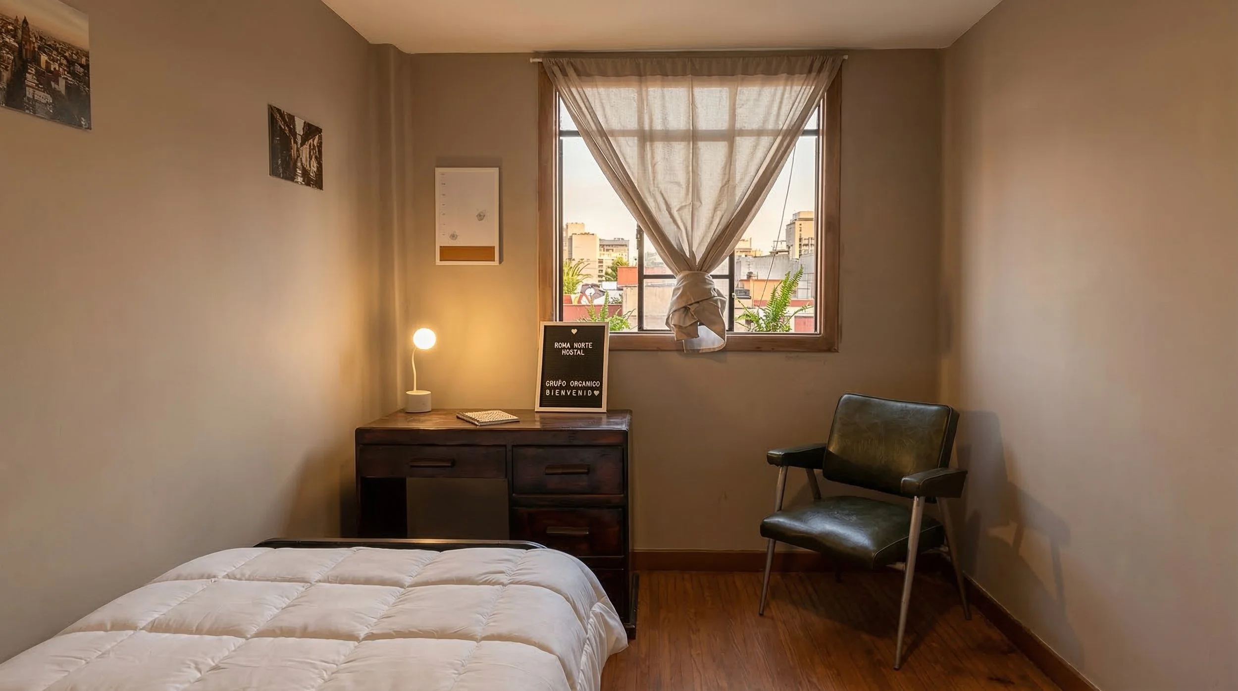 Habitación sencilla con cama, escritorio de madera, silla de cuero, cortina blanca en la ventana y una lámpara de mesa encendida.