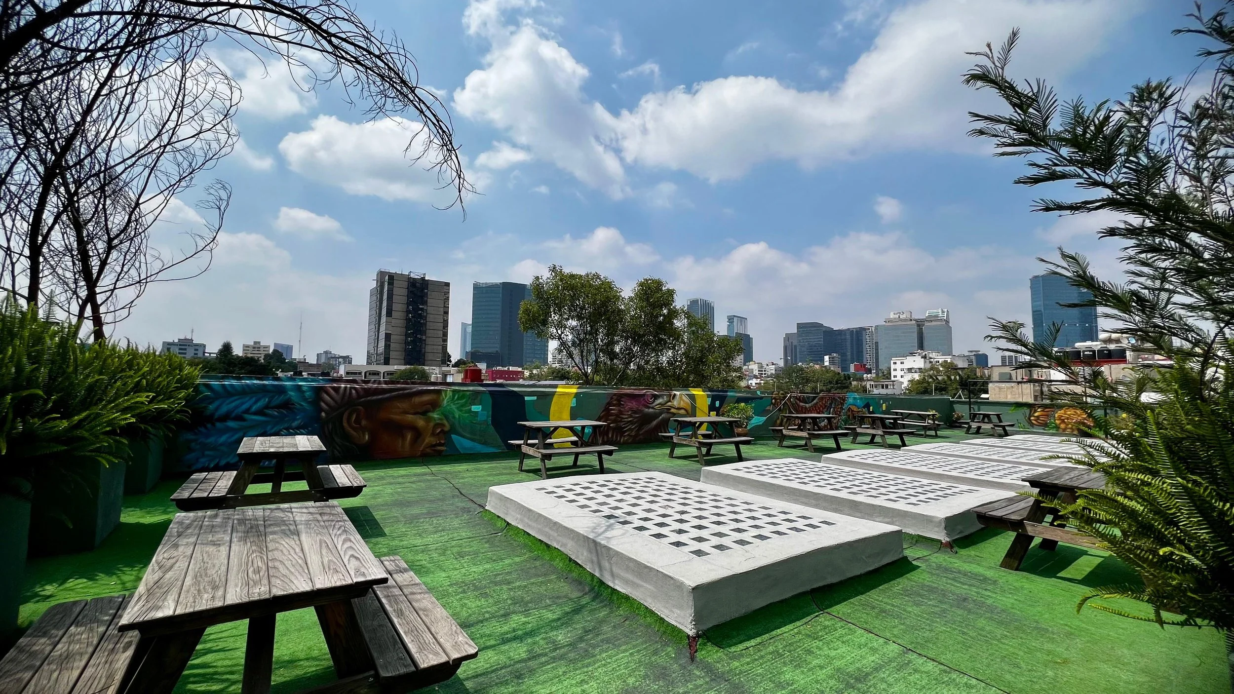 Terraza en la azotea con mesas de picnic, bancos de madera, áreas verdes y graffitis urbanos con vista a la ciudad de edificios altos y cielo con nubes.