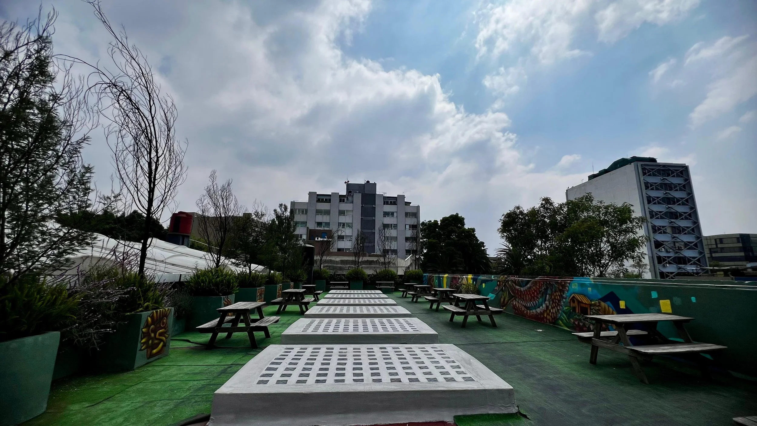 Área de descanso con mesas de madera, plantas en macetas grandes y un mural colorido en la pared, con edificios modernos y cielo nublado al fondo.