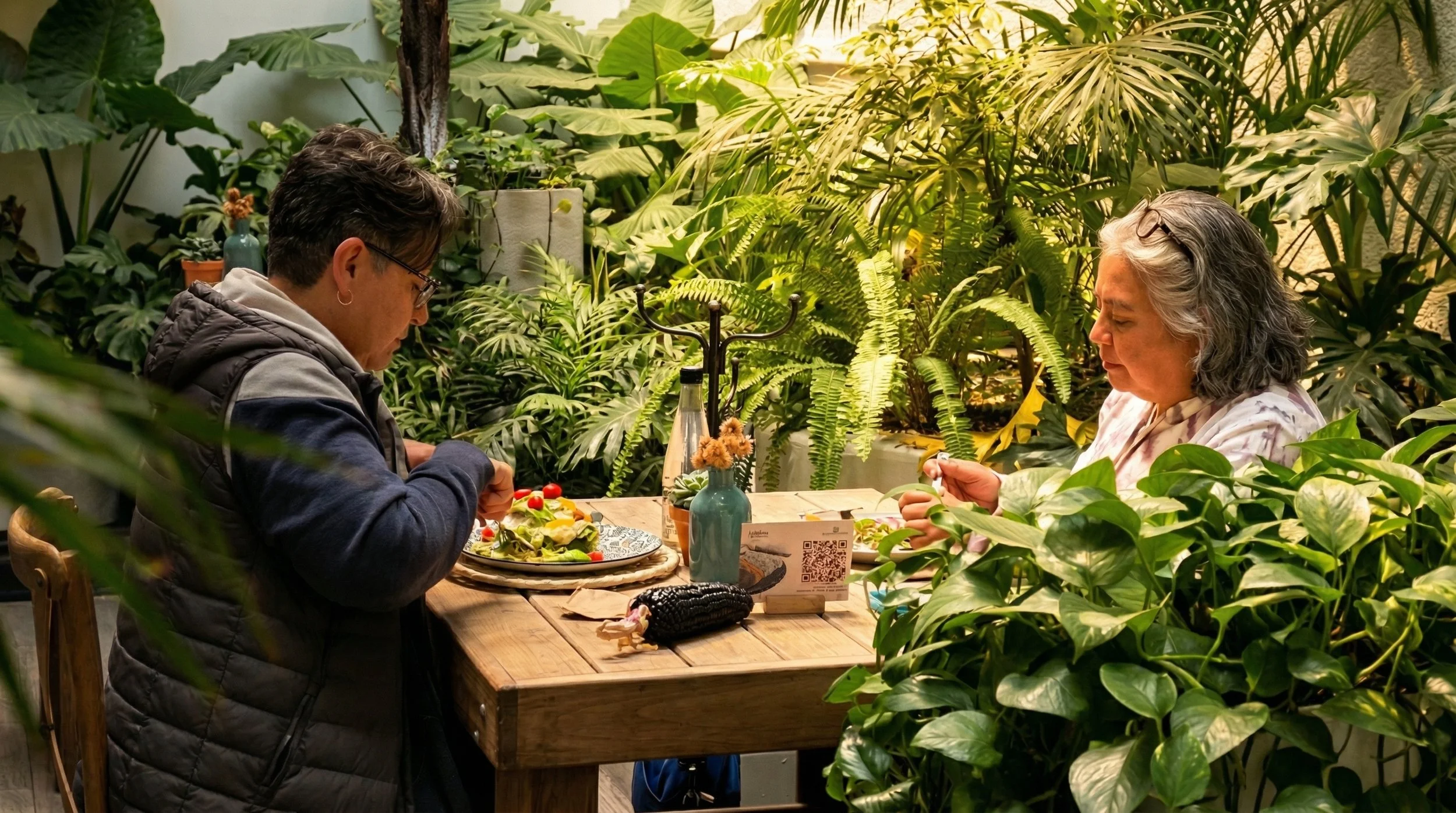 Dos personas sentadas en una mesa de madera en un ambiente lleno de plantas verdes, una mujer y un hombre, parecen estar preparando comida o trabajando en un proyecto juntos.
