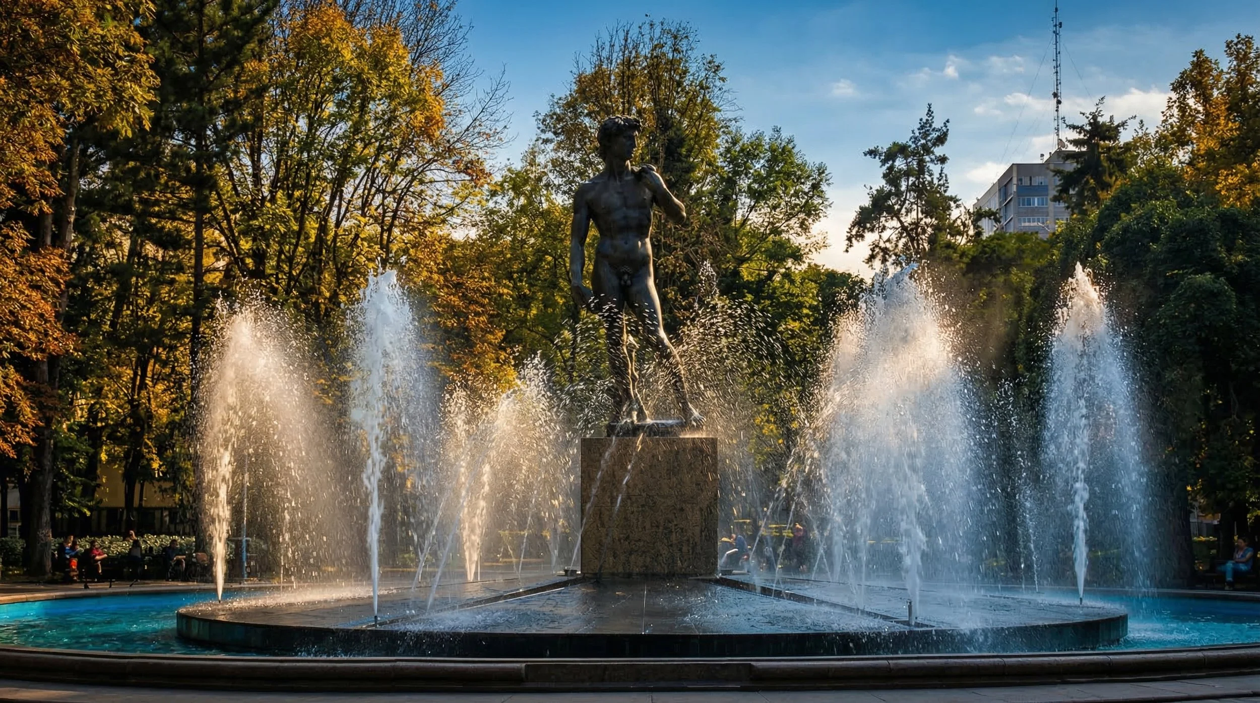 Escultura de un niño en una fuente con agua en un parque con árboles y edificios al fondo.