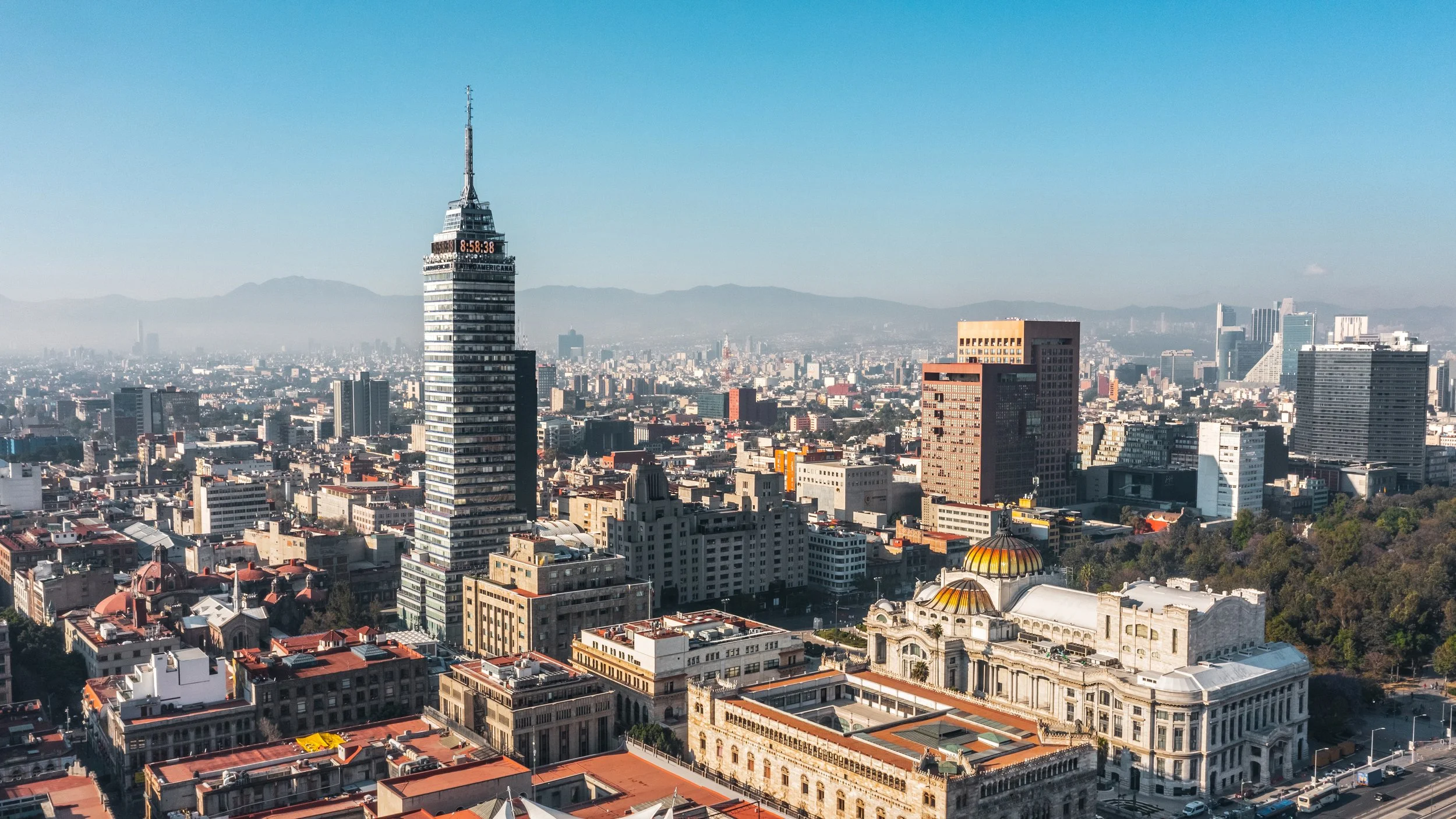 Vista panorámica de la ciudad de México, en primer plano diversos edificios históricos y modernos, incluyendo la Torre Latinoamericana, y en el fondo montañas y una ciudad extendida bajo un cielo despejado.