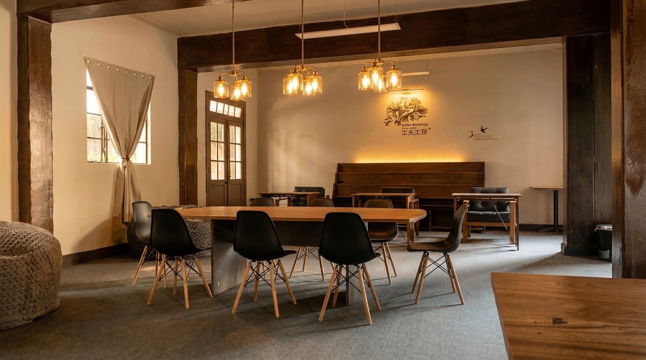 Sala de reuniones con mesas de madera y sillas negras, lámparas colgantes de vidrio, ventanas con cortinas beige, pared con arte y logotipo de un árbol, ambiente cálido y acogedor.