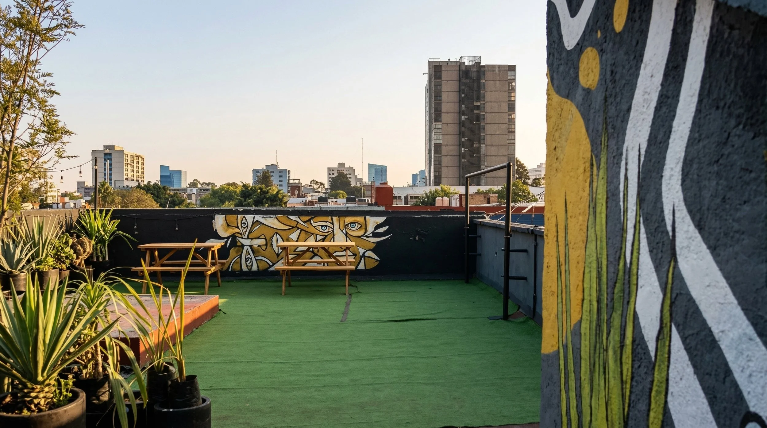 Terraza en la ciudad con mesas de picnic, plantas en macetas y grafiti en la pared de fondo. Edificios y árboles en el horizonte.