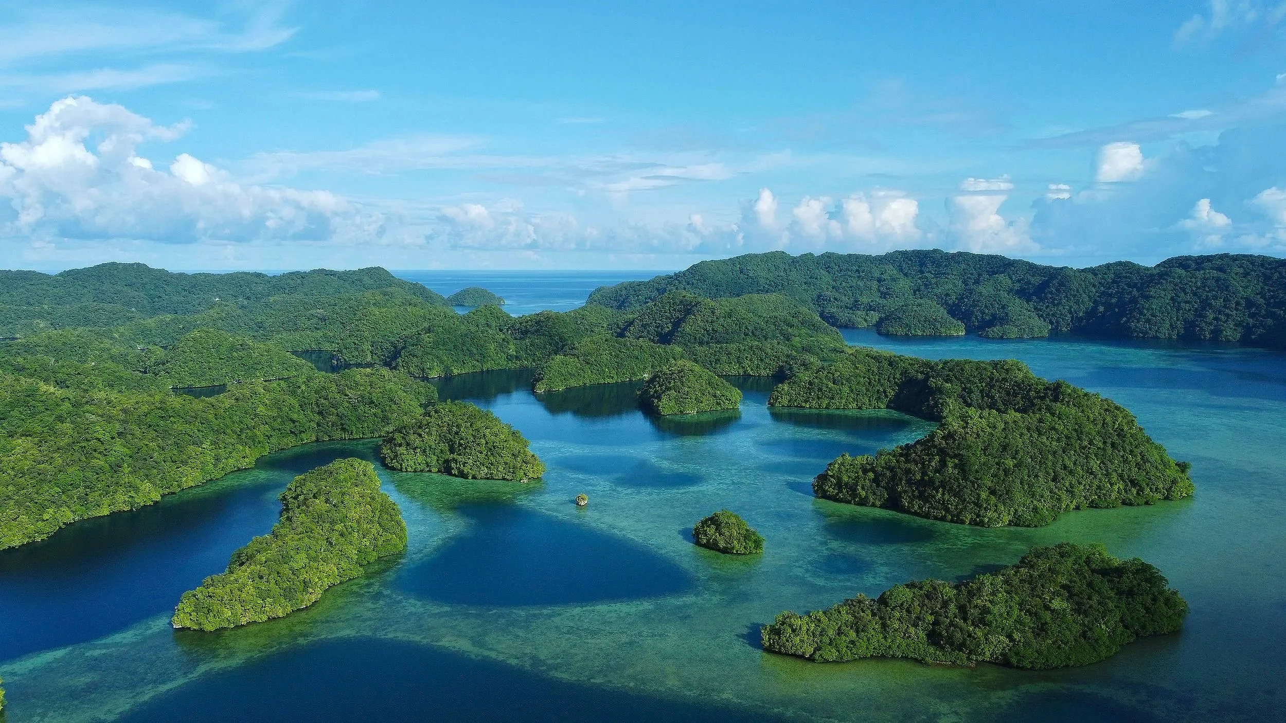 Aerial view of a lush green island with numerous small islands and inlets surrounded by turquoise blue water, under a partly cloudy sky.