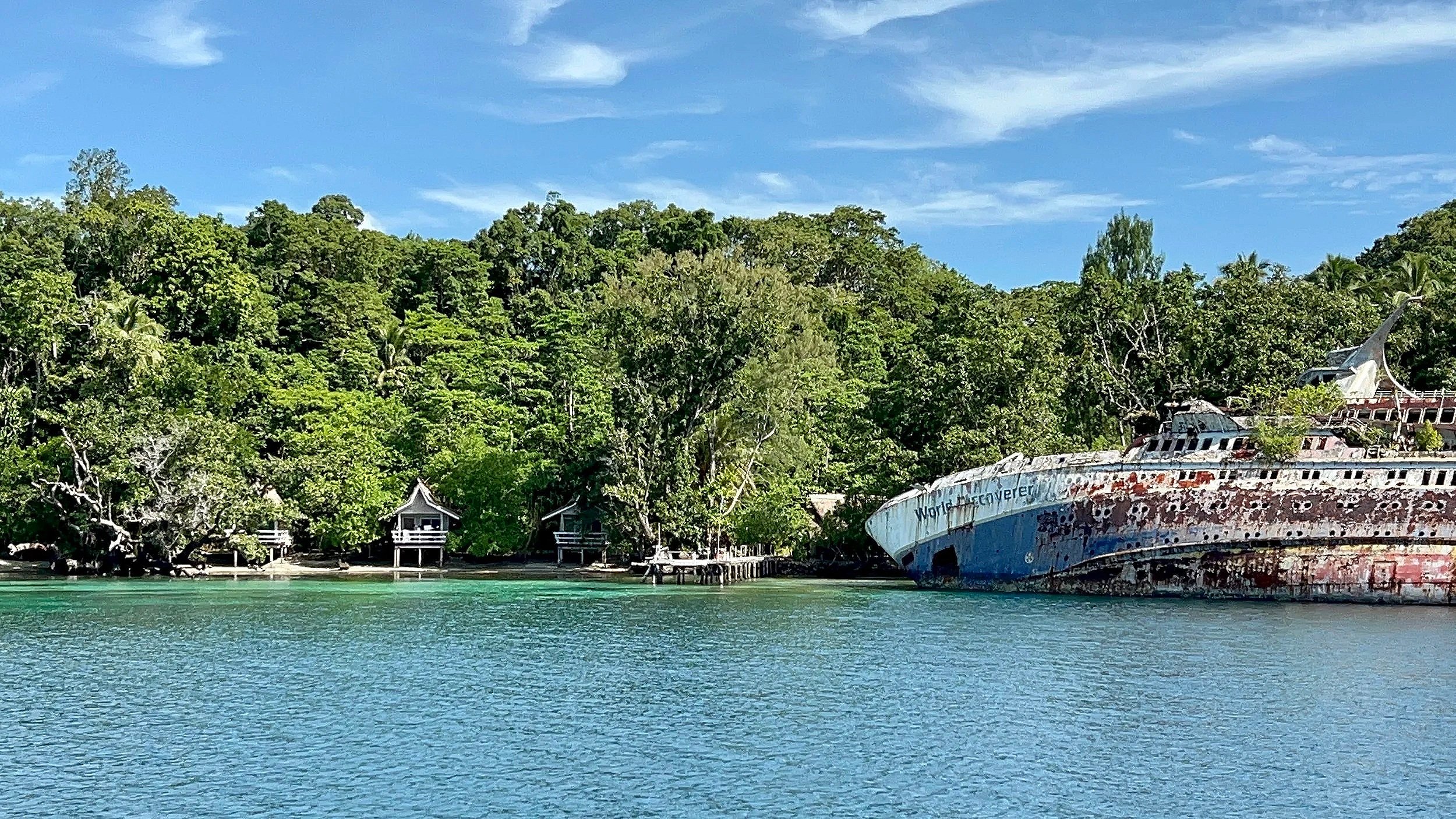 An abandoned, rusted ship near the shore with lush green trees and small pavilions along the water's edge on a sunny day.
