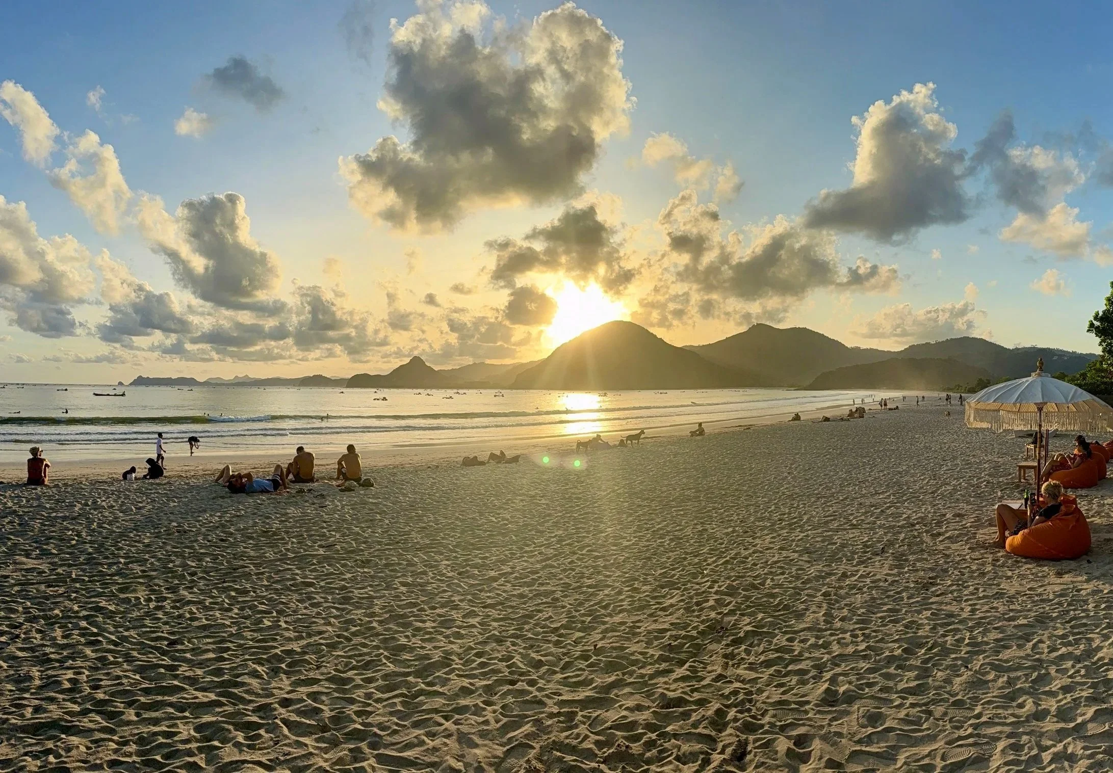 A beach scene at sunset with people relaxing on the sand, some under umbrellas, others lying or sitting in small groups. The ocean is calm with a few boats visible, and there are mountains in the distance with a partly cloudy sky.