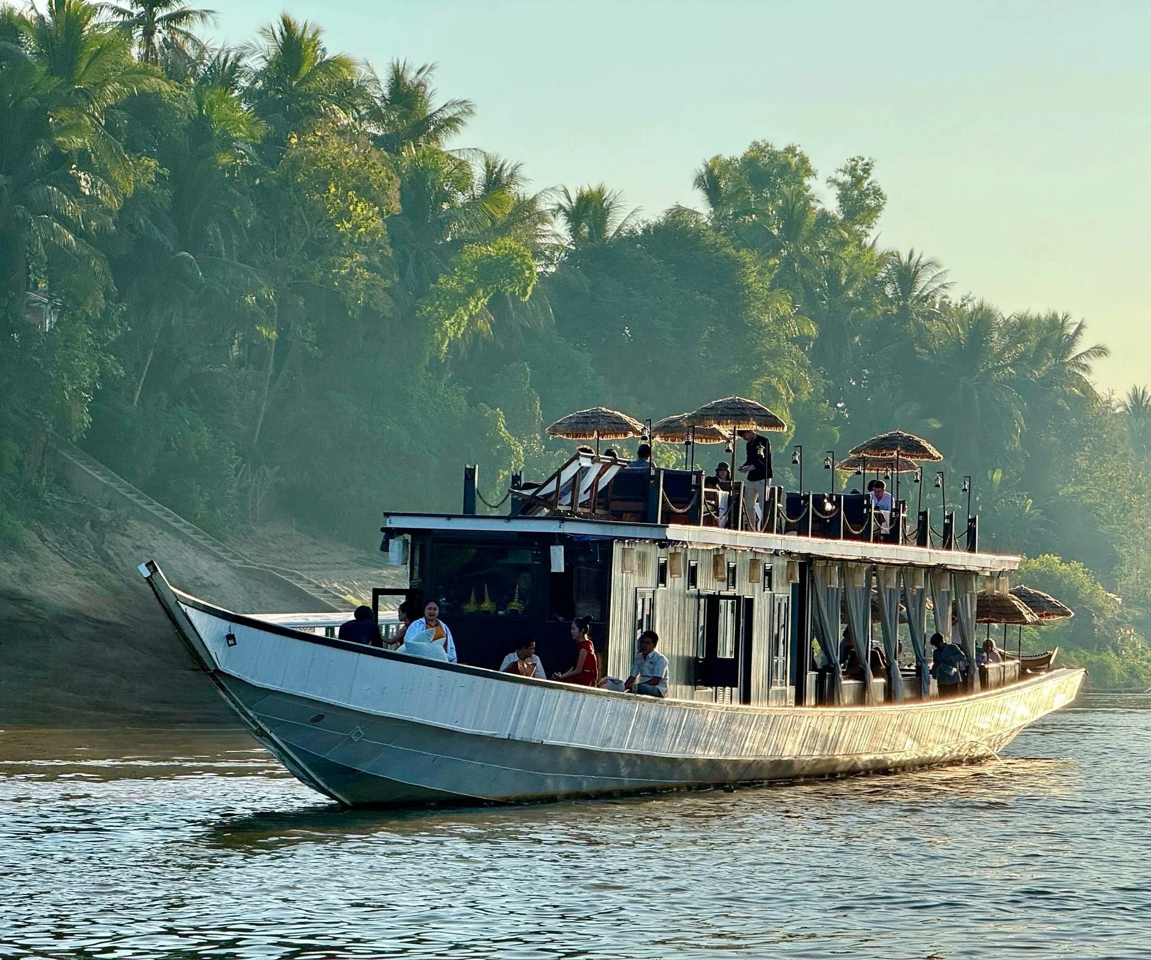 A boat with people on board sailing on a river surrounded by lush green trees, some people seated and standing on the boat's lower and upper decks.
