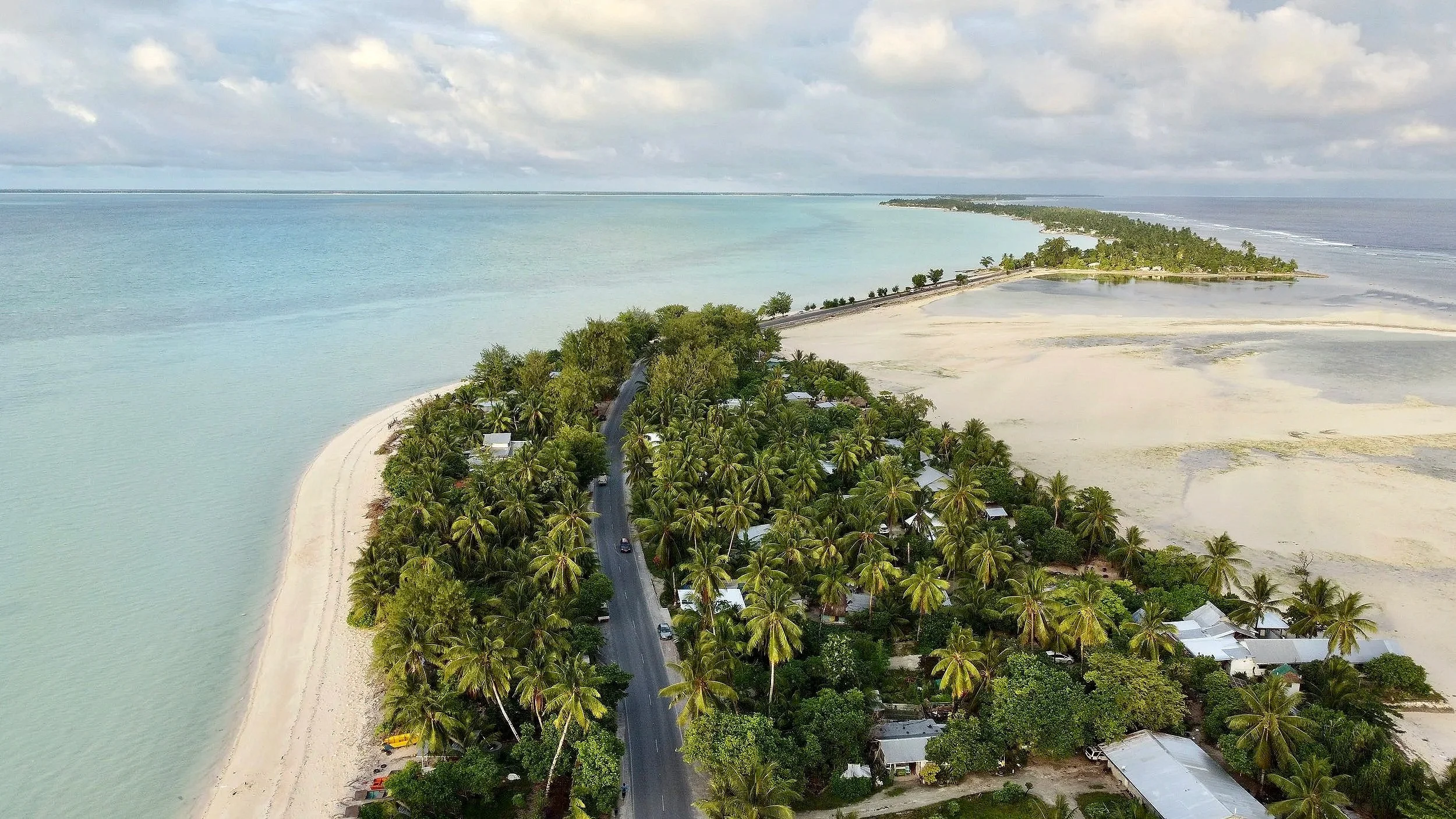 Aerial view of a tropical island with a road running through the dense green palm trees and small buildings, surrounded by ocean and sandy beaches.