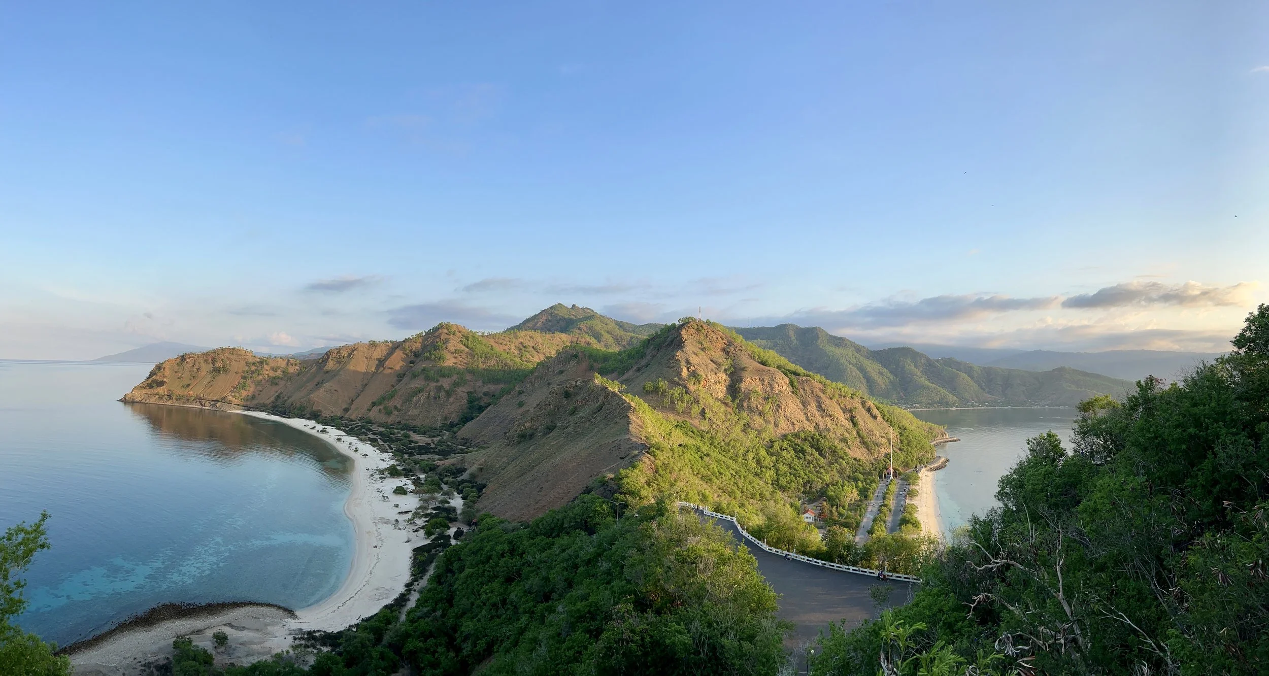 Scenic view of a coastal landscape with green hills, a sandy beach, and calm water under a blue sky with some clouds.