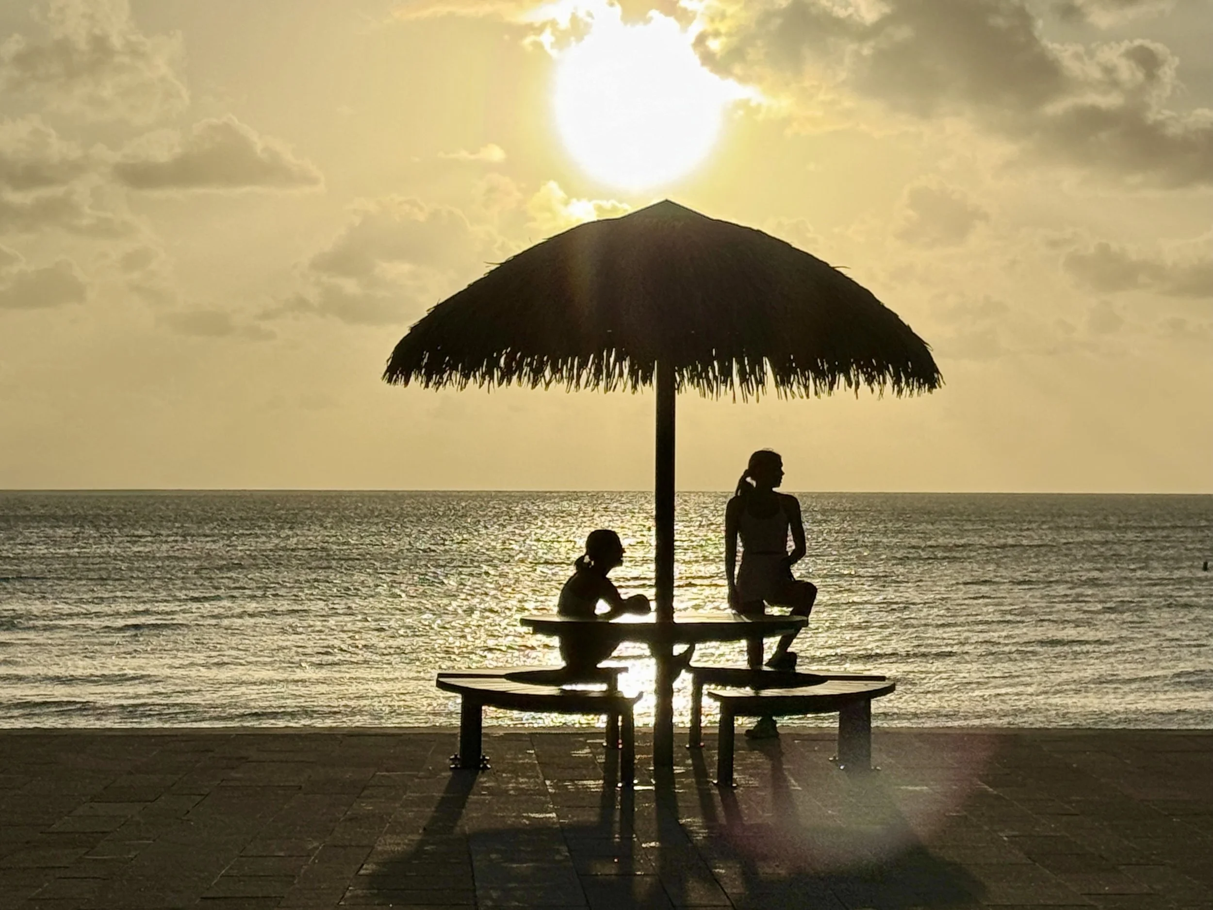 Silhouettes of two women under a beach umbrella on a seaside promenade at sunset, with the ocean and cloudy sky in the background.