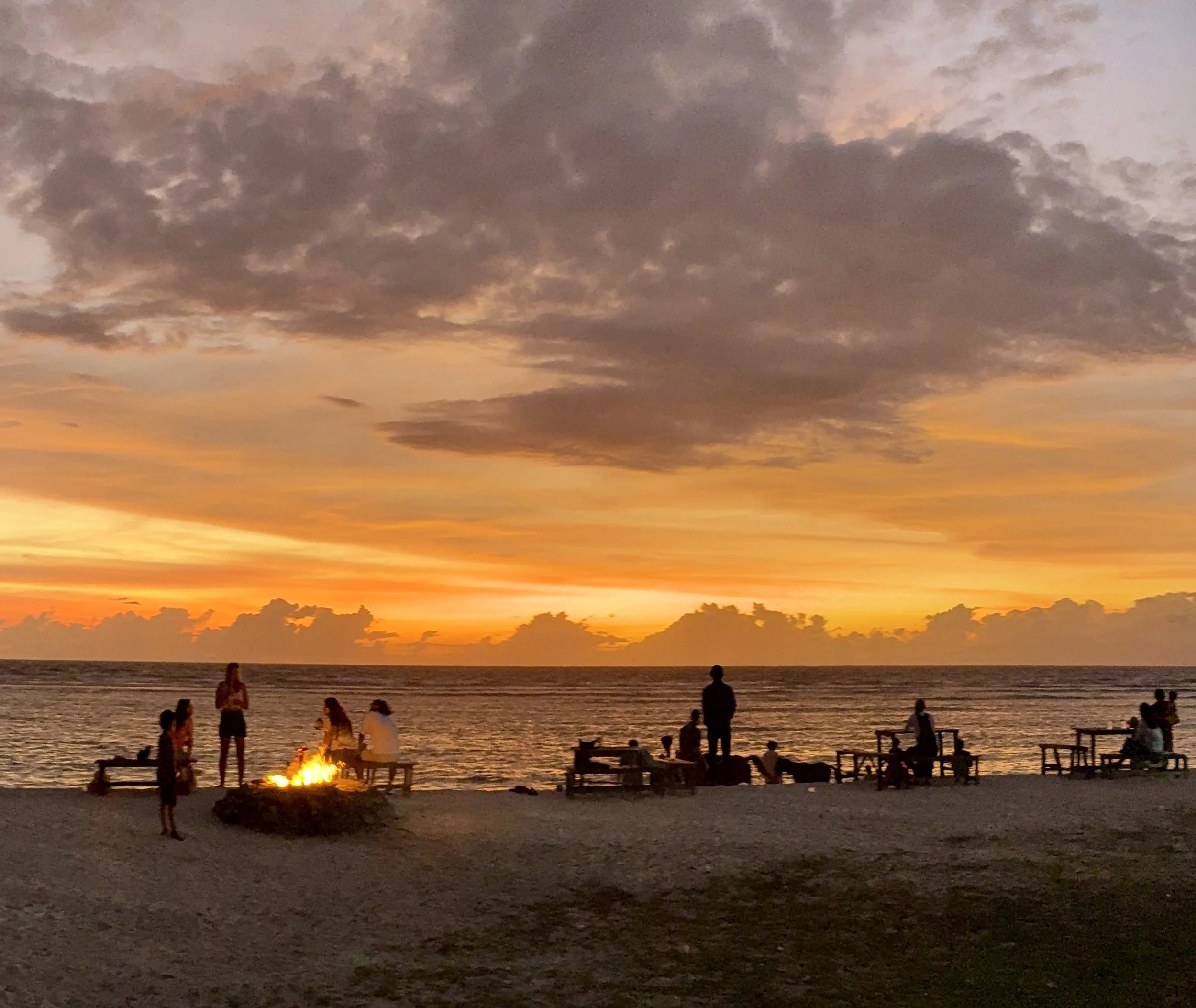 People gathered around a fire on a beach during sunset with colorful clouds in the sky.