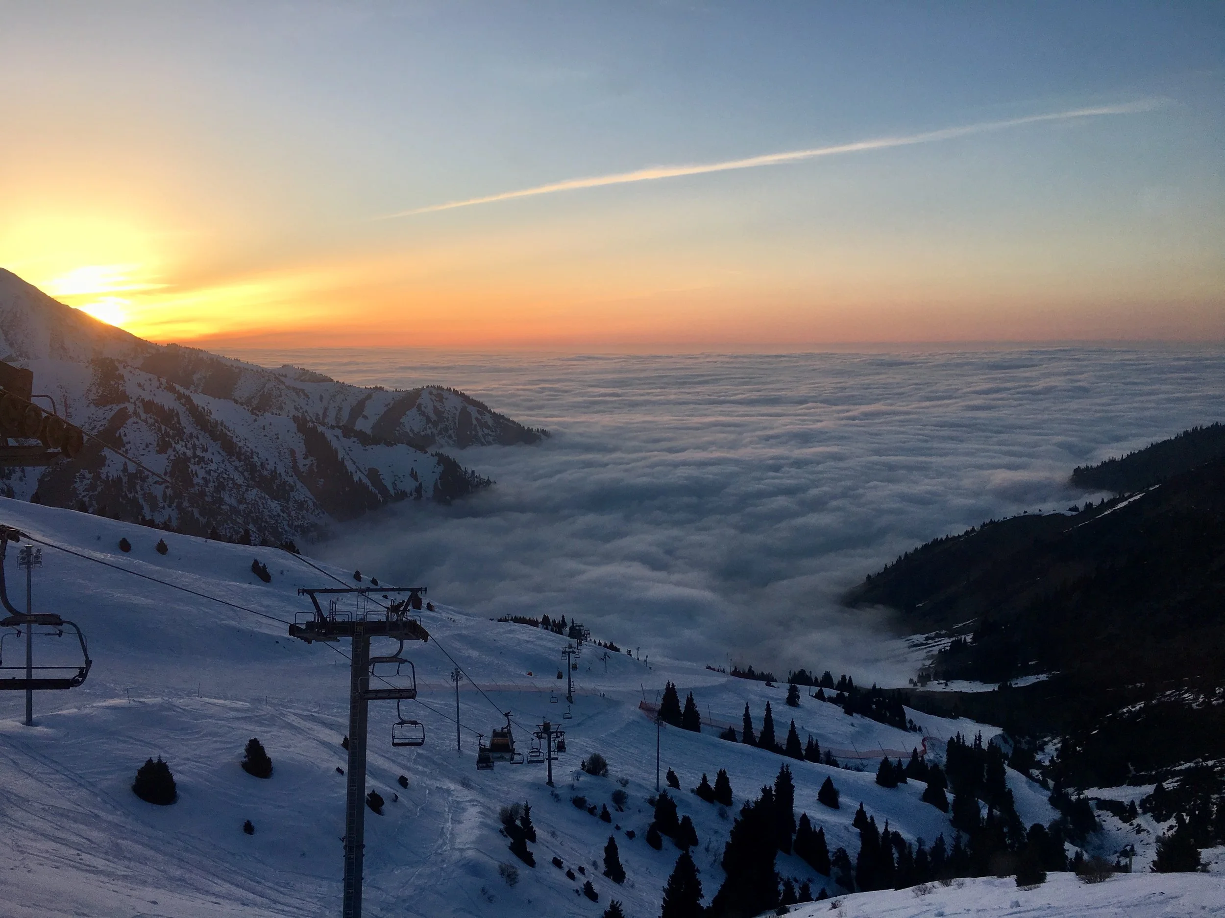 Sunset over snow-covered mountains with ski lift chairs in the foreground, and a blanket of clouds in the valley below.