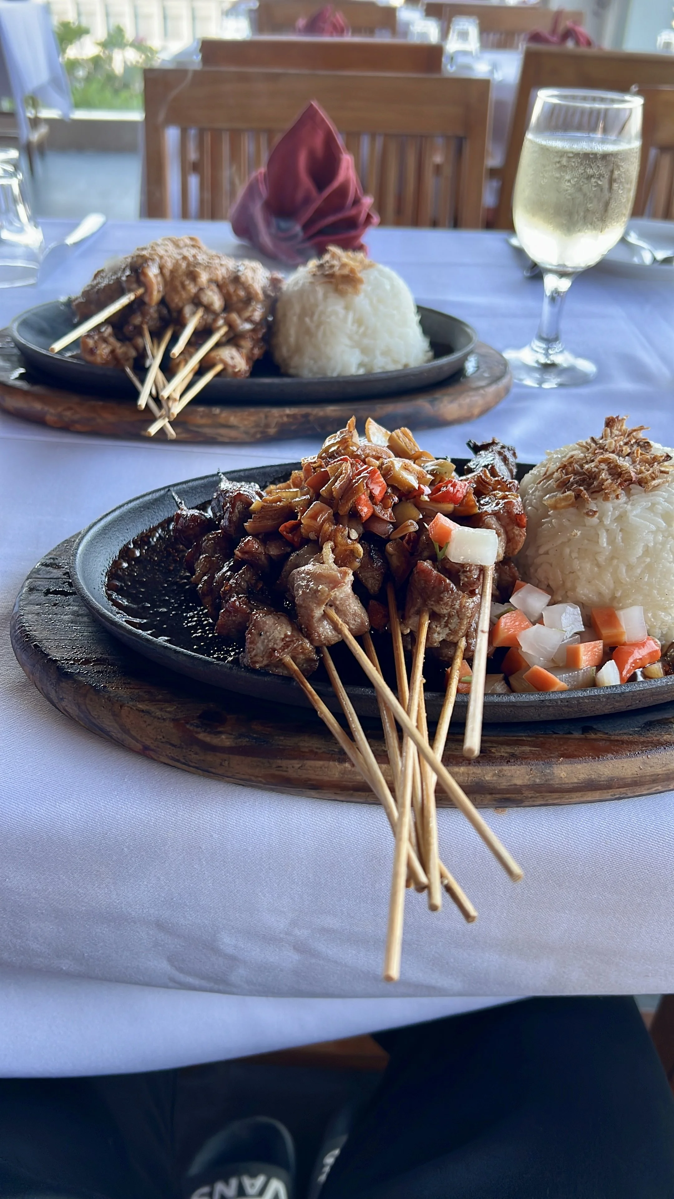 Two sizzling platters of skewered meat with rice and vegetables, one with a side of pickled vegetables, on a white tablecloth in a restaurant setting with a glass of white wine.