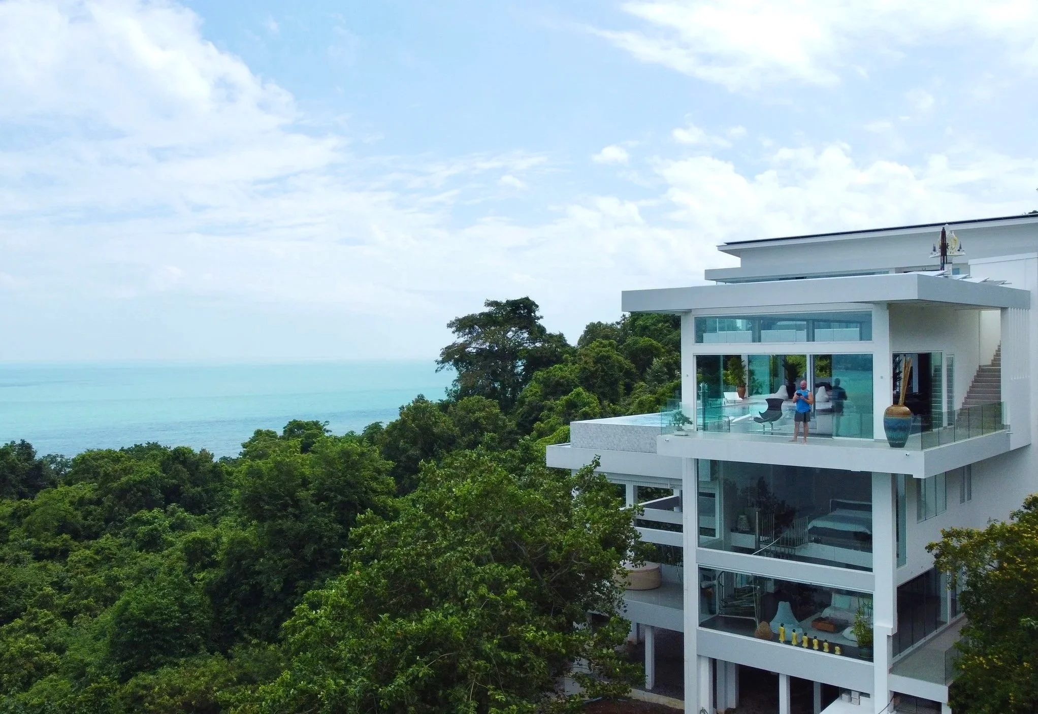 Modern white house with large glass windows overlooking a lush green forest and the ocean in the background.