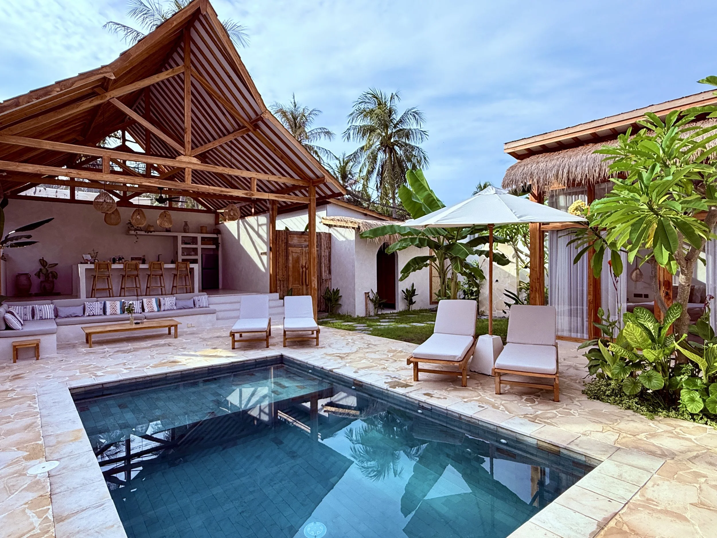 Luxury outdoor pool area with lounge chairs, umbrellas, tropical plants, and a covered patio with a bar