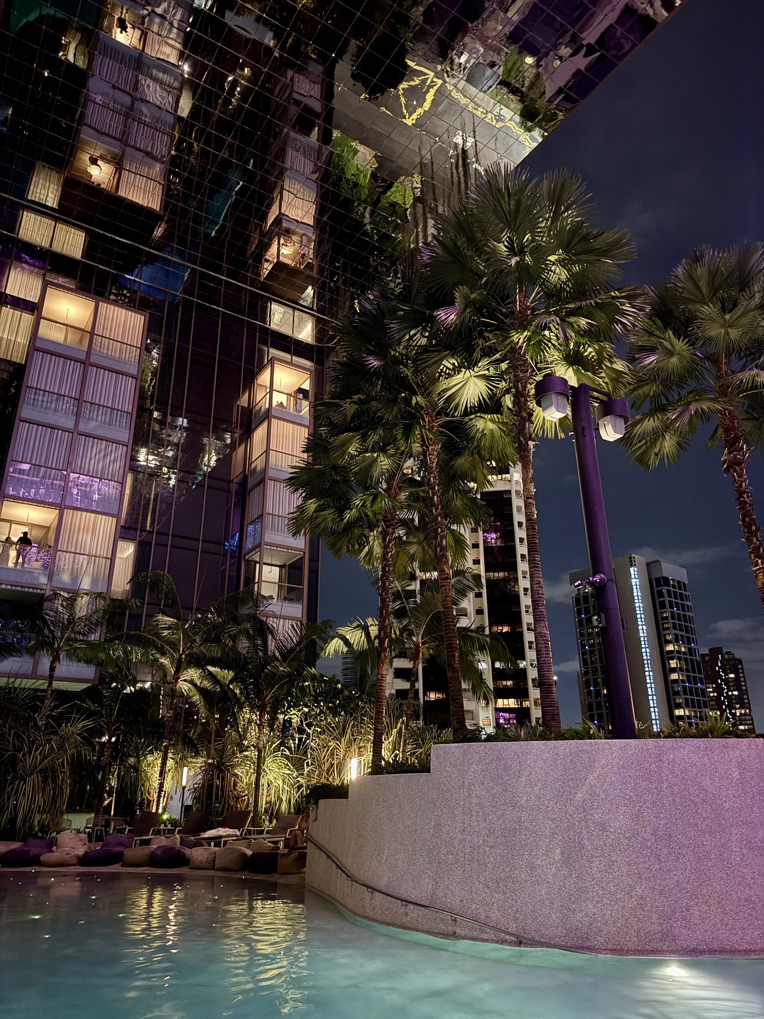 Nighttime outdoor scene with a swimming pool, surrounded by palm trees, lounge chairs, and tall modern buildings with illuminated windows and balconies.