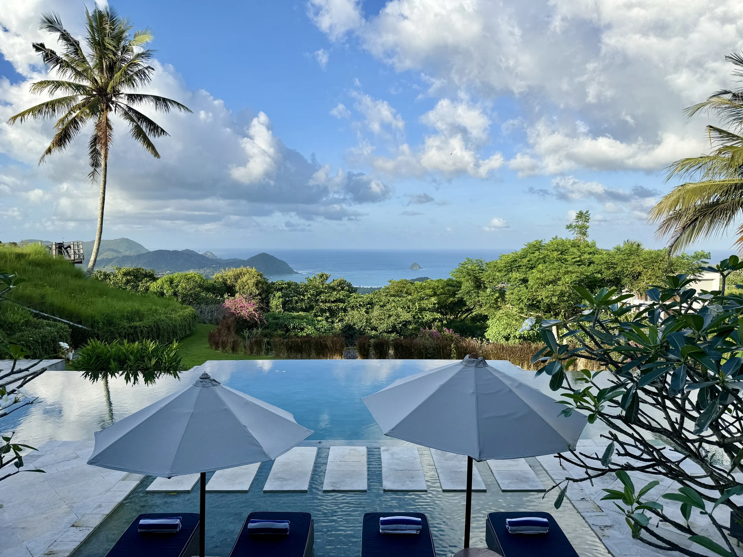 View of a swimming pool with two lounge chairs and umbrellas, overlooking lush green gardens and the ocean in the distance on a sunny day with partly cloudy skies.