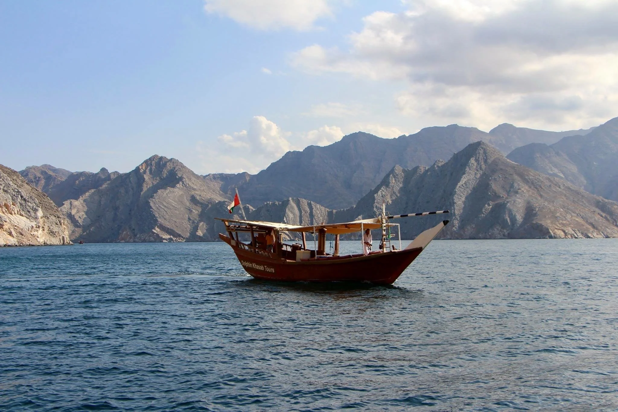 A wooden boat with a flag on the water, surrounded by mountainous terrain and partly cloudy sky.