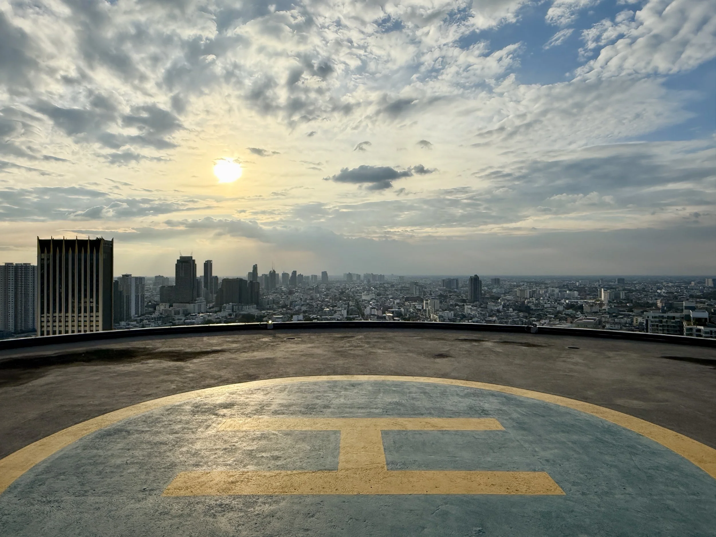 View of a city skyline at sunset or sunrise, seen from a helicopter landing pad on top of a building, with the city stretching to the horizon under a partly cloudy sky.