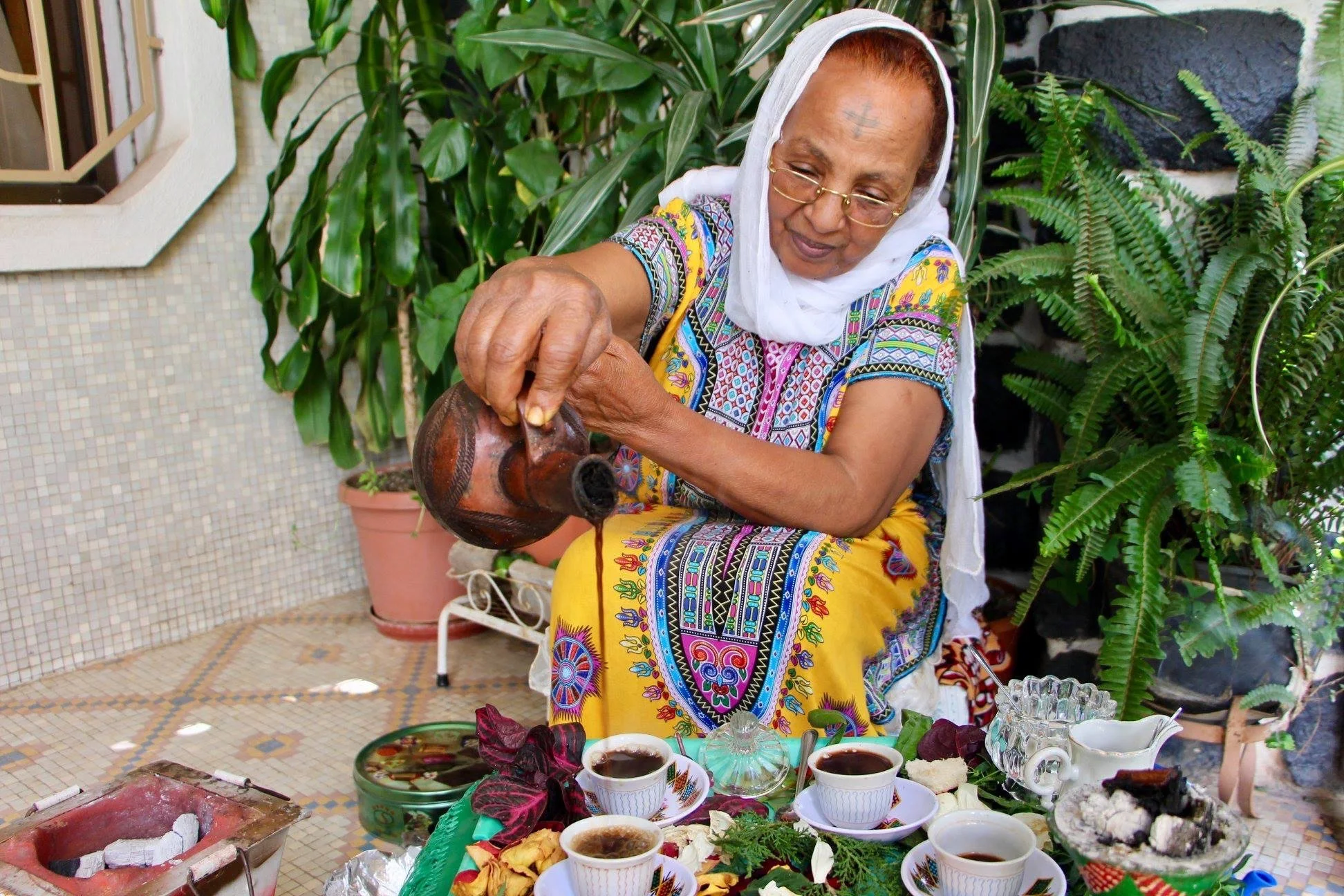 An elderly woman dressed in colorful traditional clothing pouring tea into cups on a table with plants and potted greenery in the background.