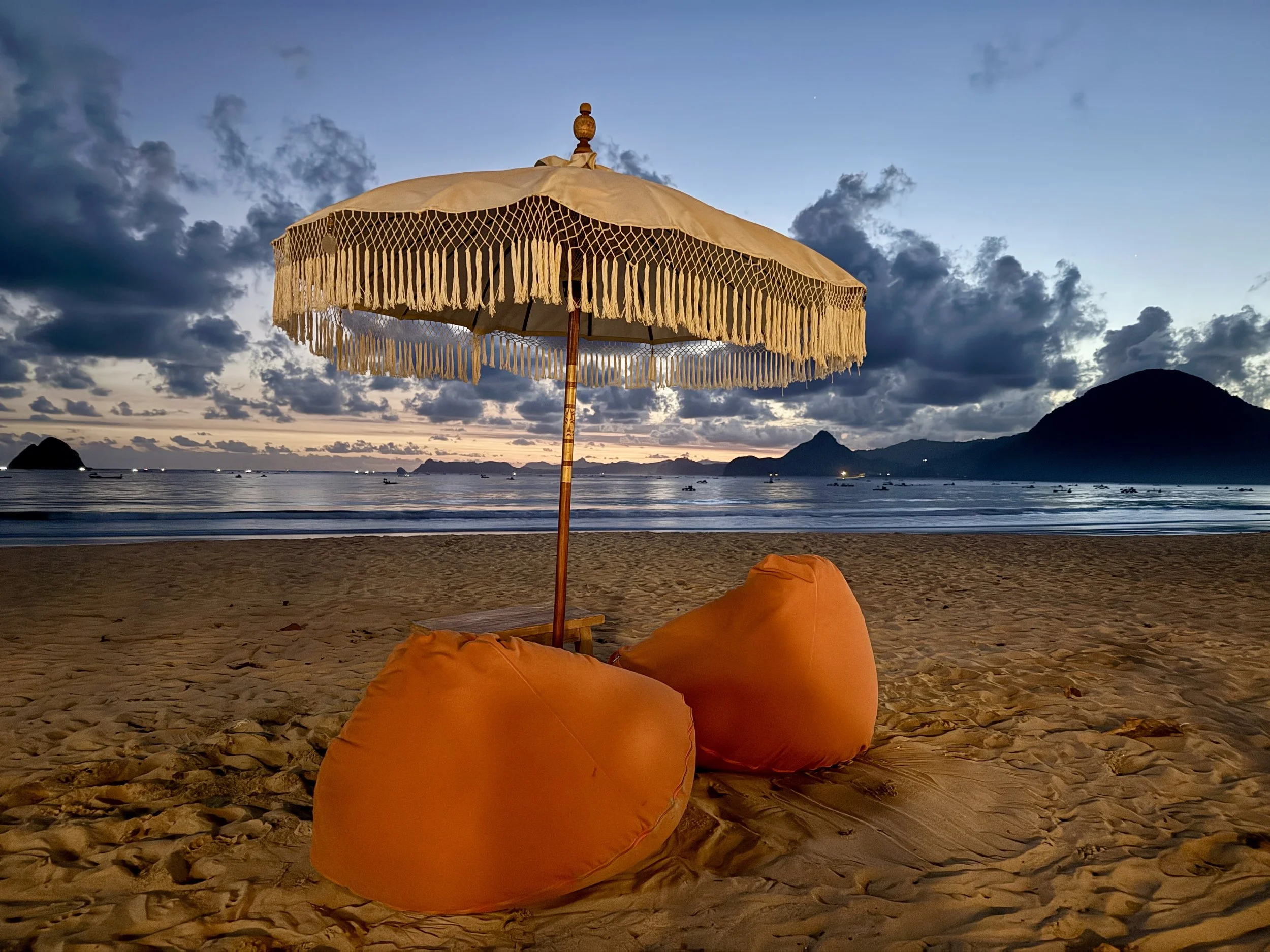 Beach scene at sunset with a large cream-colored umbrella with fringes, two orange bean bags on the sand, calm ocean water, boats, and distant mountains under a partly cloudy sky.