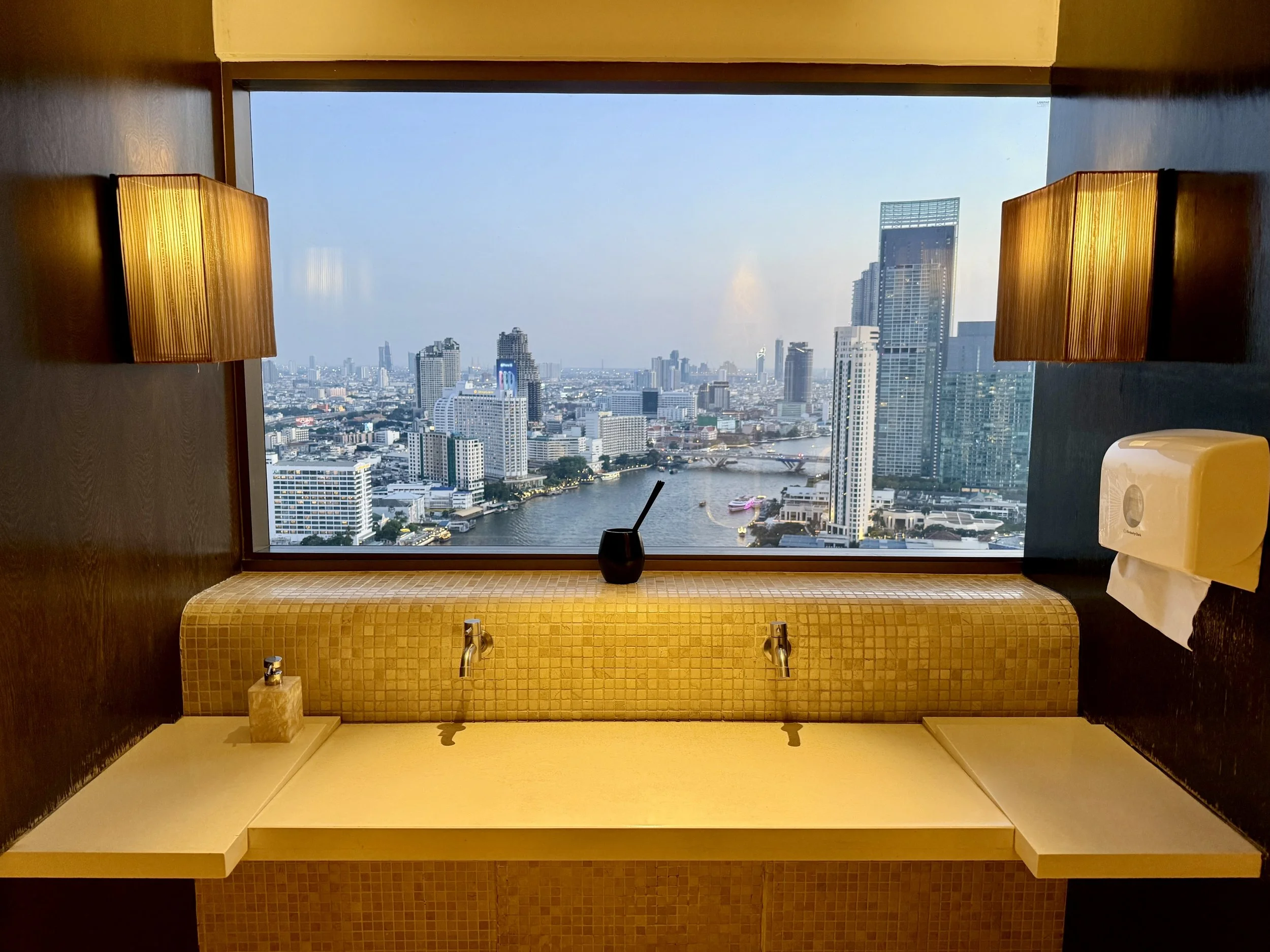 City view of high-rise buildings and a river seen through a large window, with a bathroom sink and mirror in the foreground.