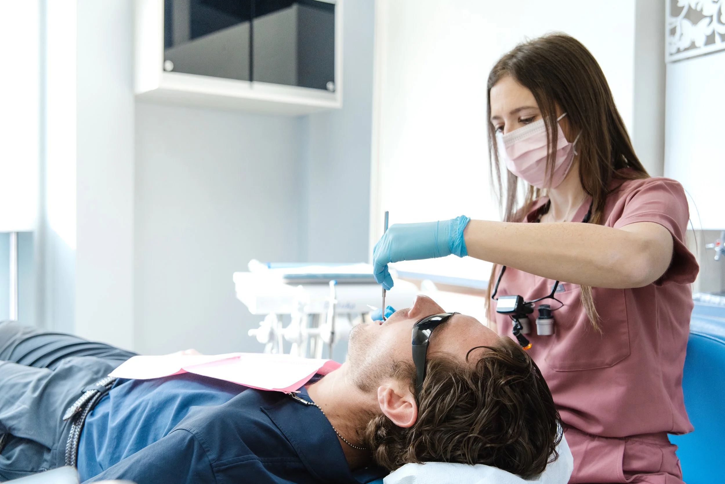 A dental hygienist in pink scrubs and face mask performing dental work on a patient lying in a dental chair, with the patient's mouth open and wearing safety glasses, in a dental clinic.
