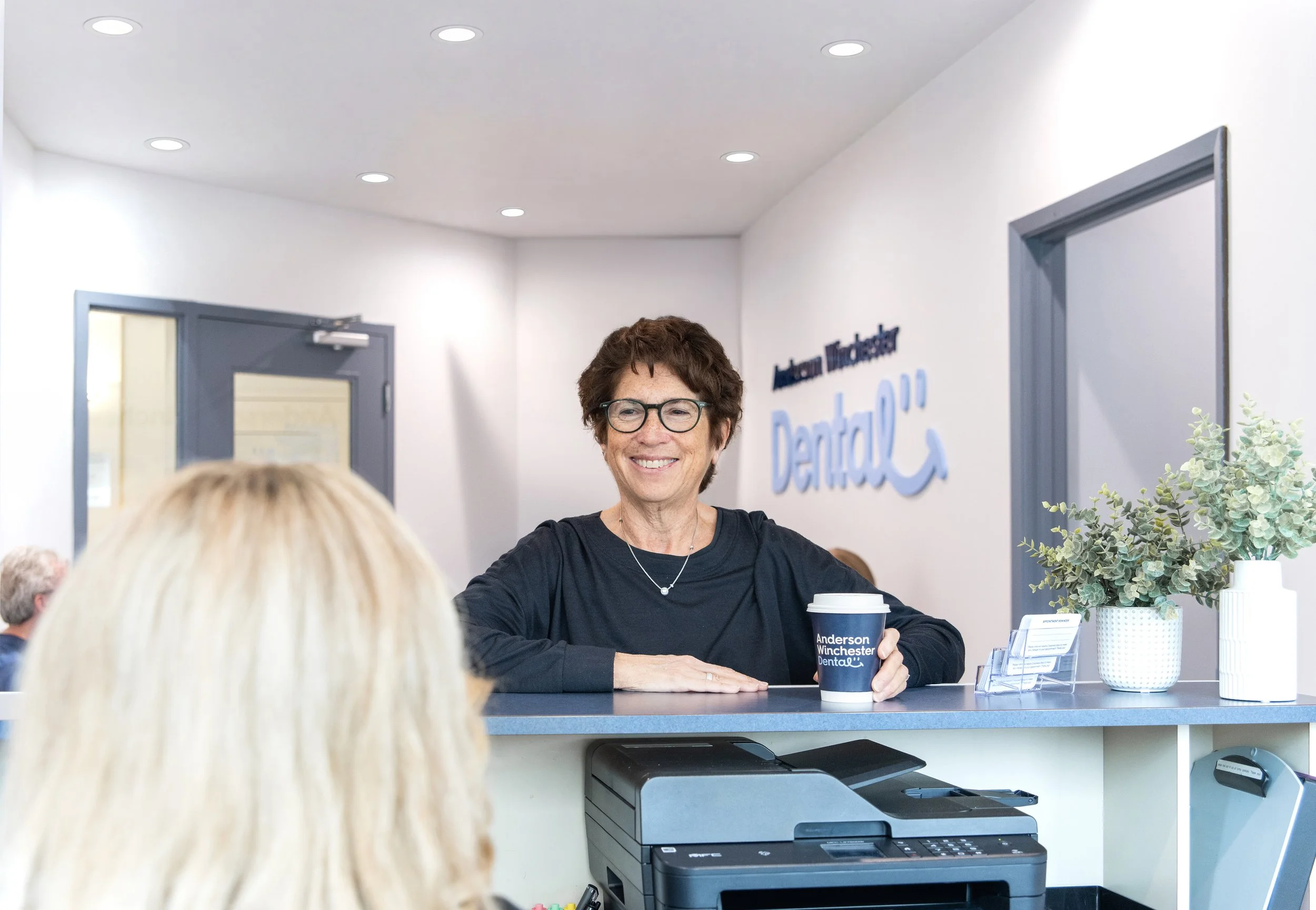 A woman with short brown hair and glasses smiling while sitting at a reception desk, holding a cup in her right hand labeled "Anderson Winchester Dental." The background shows a white wall with a sign that reads "Anderson Winchester Dental," a doorway, and a potted plant on the desk.