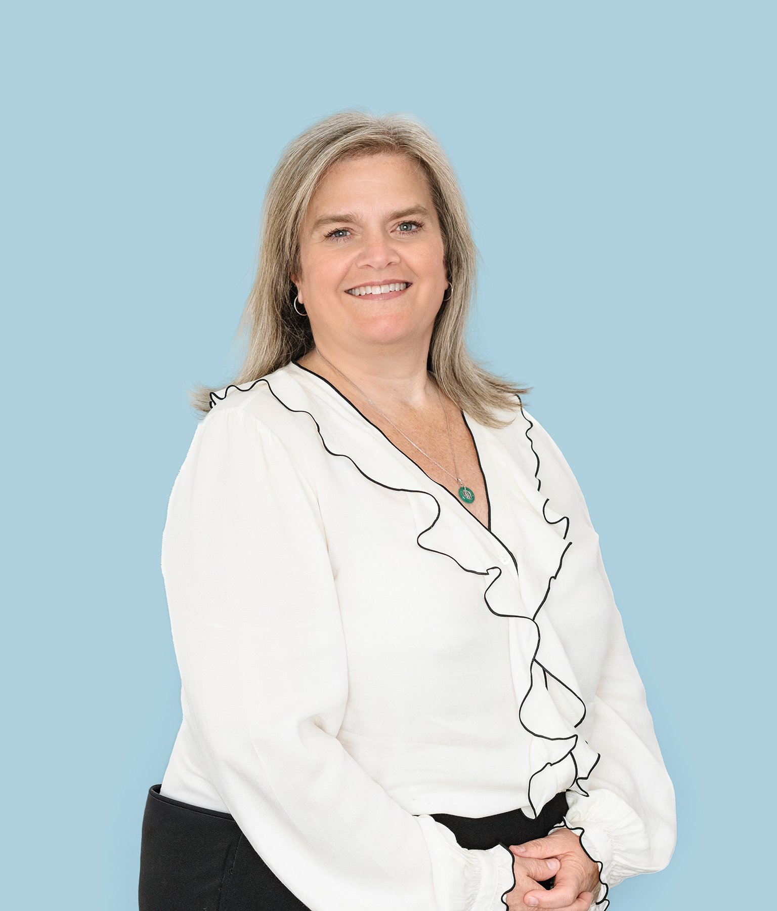 A woman with blonde hair smiling at the camera, wearing a white blouse with black ruffle trim and black pants, standing against a light blue background.