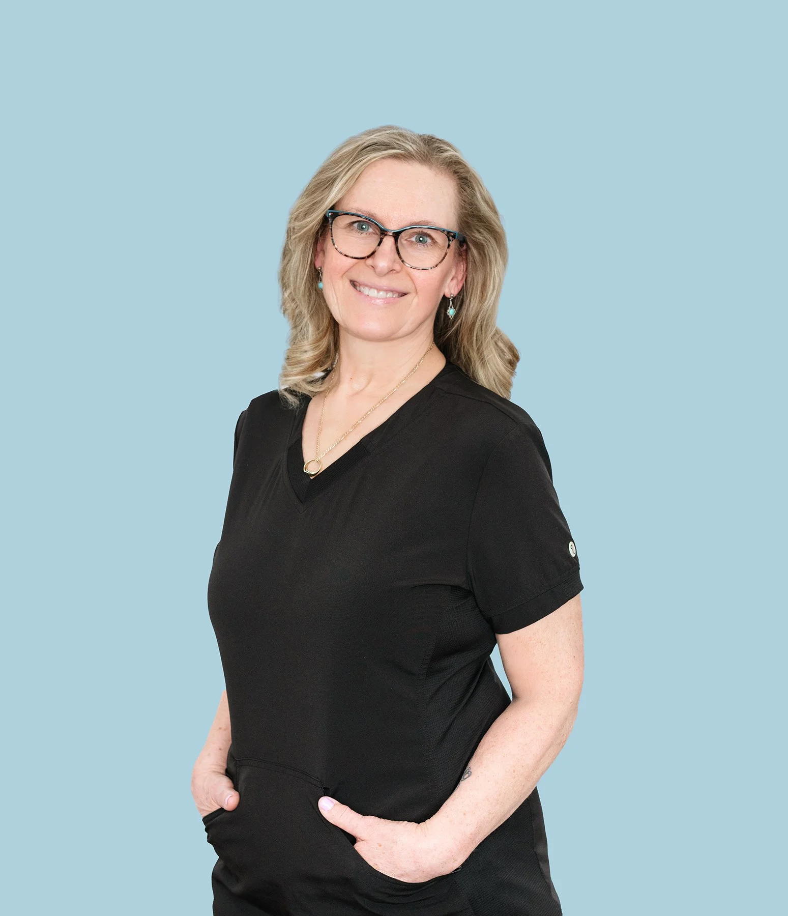 Smiling woman with blonde hair, glasses, and turquoise earrings wearing a black top against a light blue background.