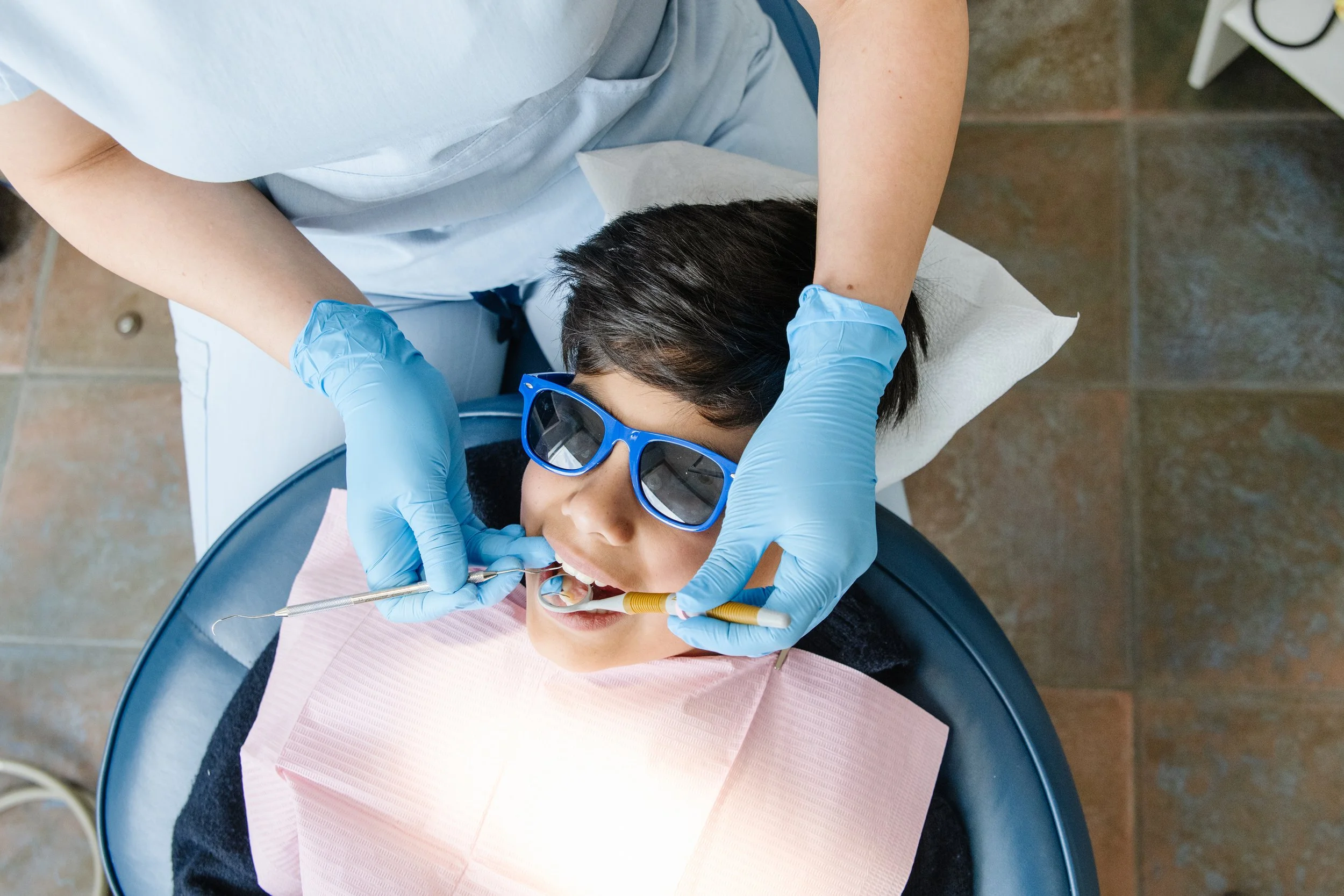 A woman lying in a dental chair with protective sunglasses, having her teeth examined or treated by a dental professional wearing blue gloves and scrubs.