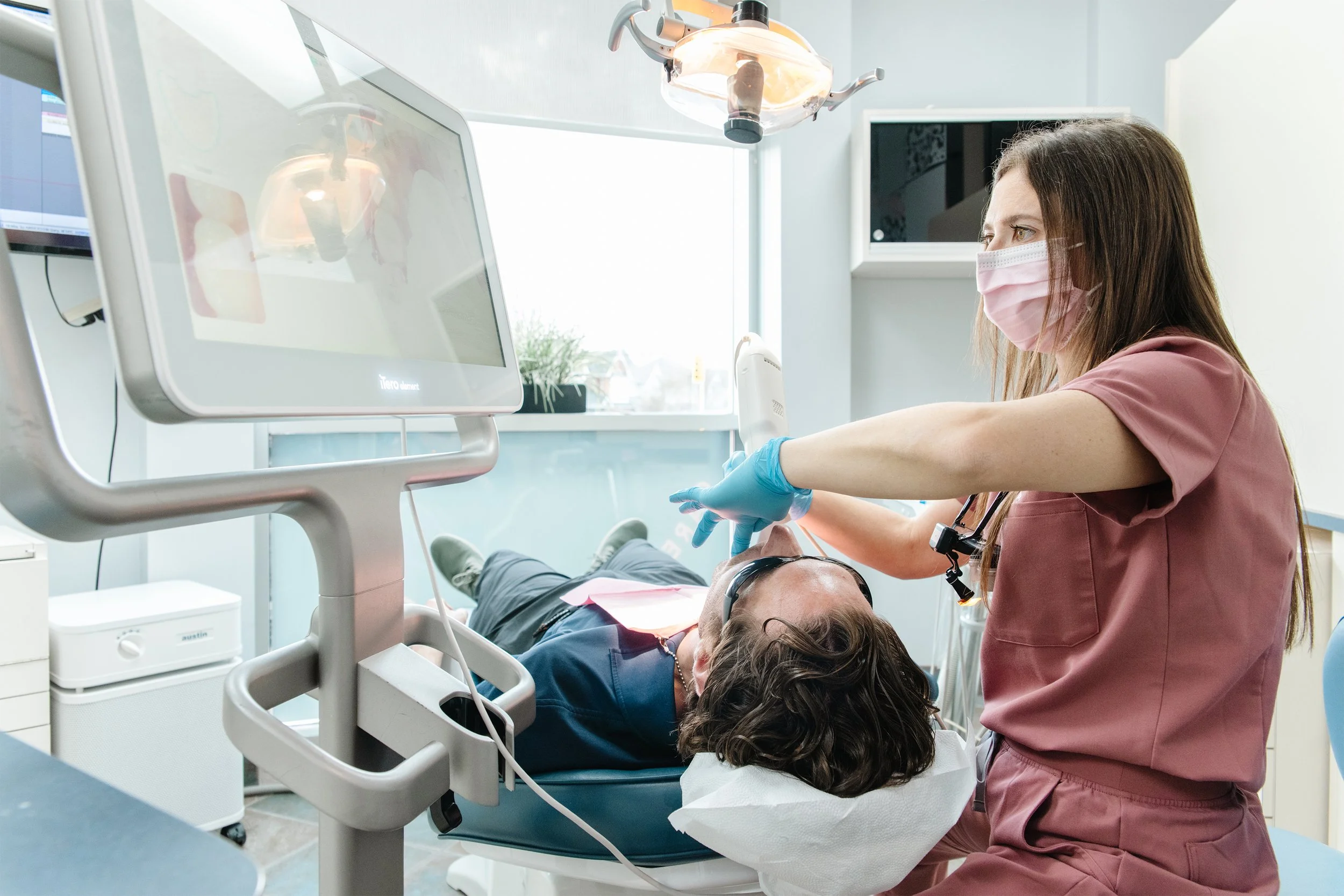 A healthcare professional in pink scrubs and a face mask is administering a dental or medical procedure to a patient lying on a treatment chair. There are medical monitors, equipment, and an overhead light in the room.