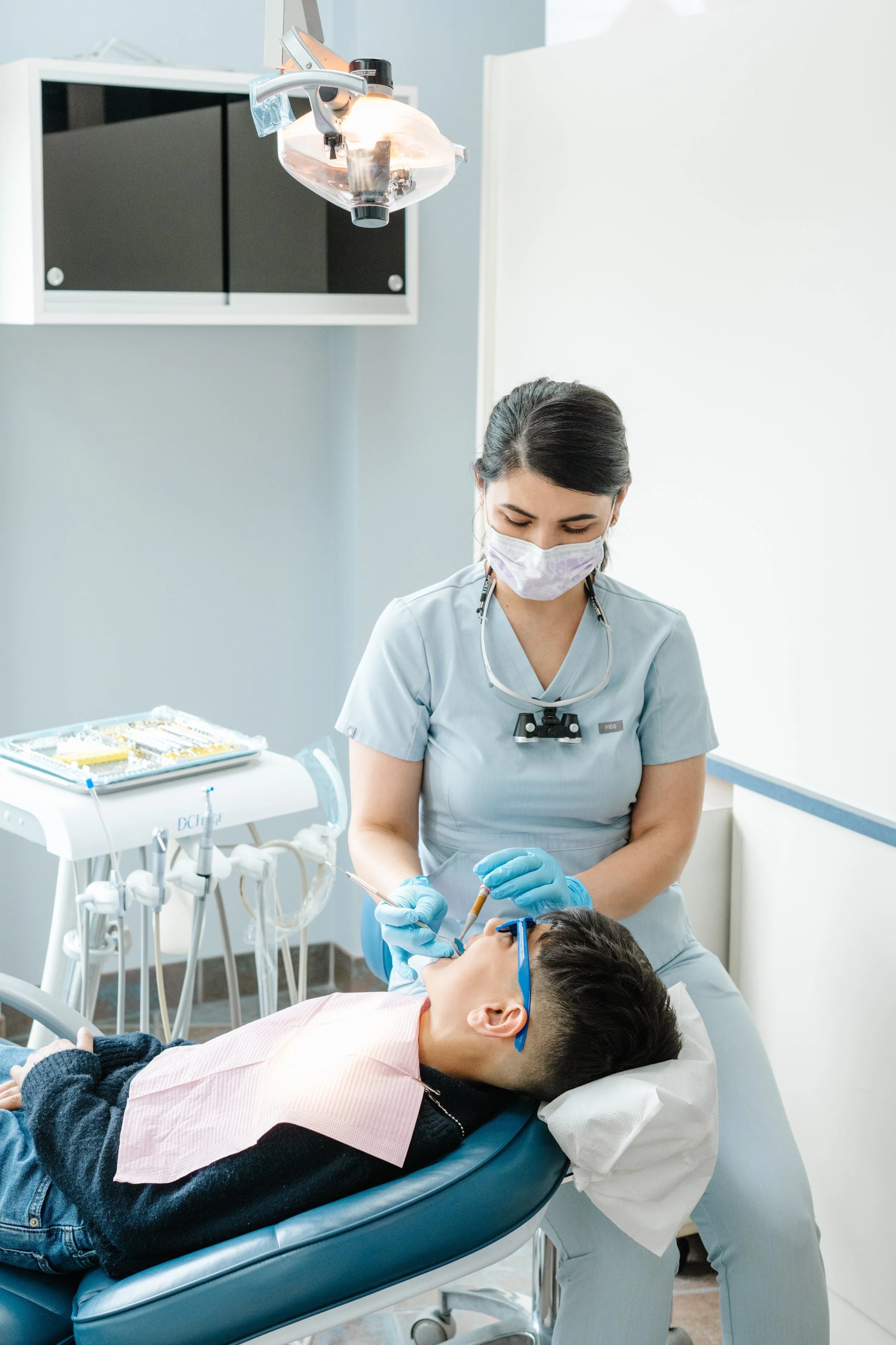 A dental assistant in scrubs and a face mask preparing to treat a patient lying in a dental chair. The patient is wearing protective glasses, and dental equipment is visible in the background.