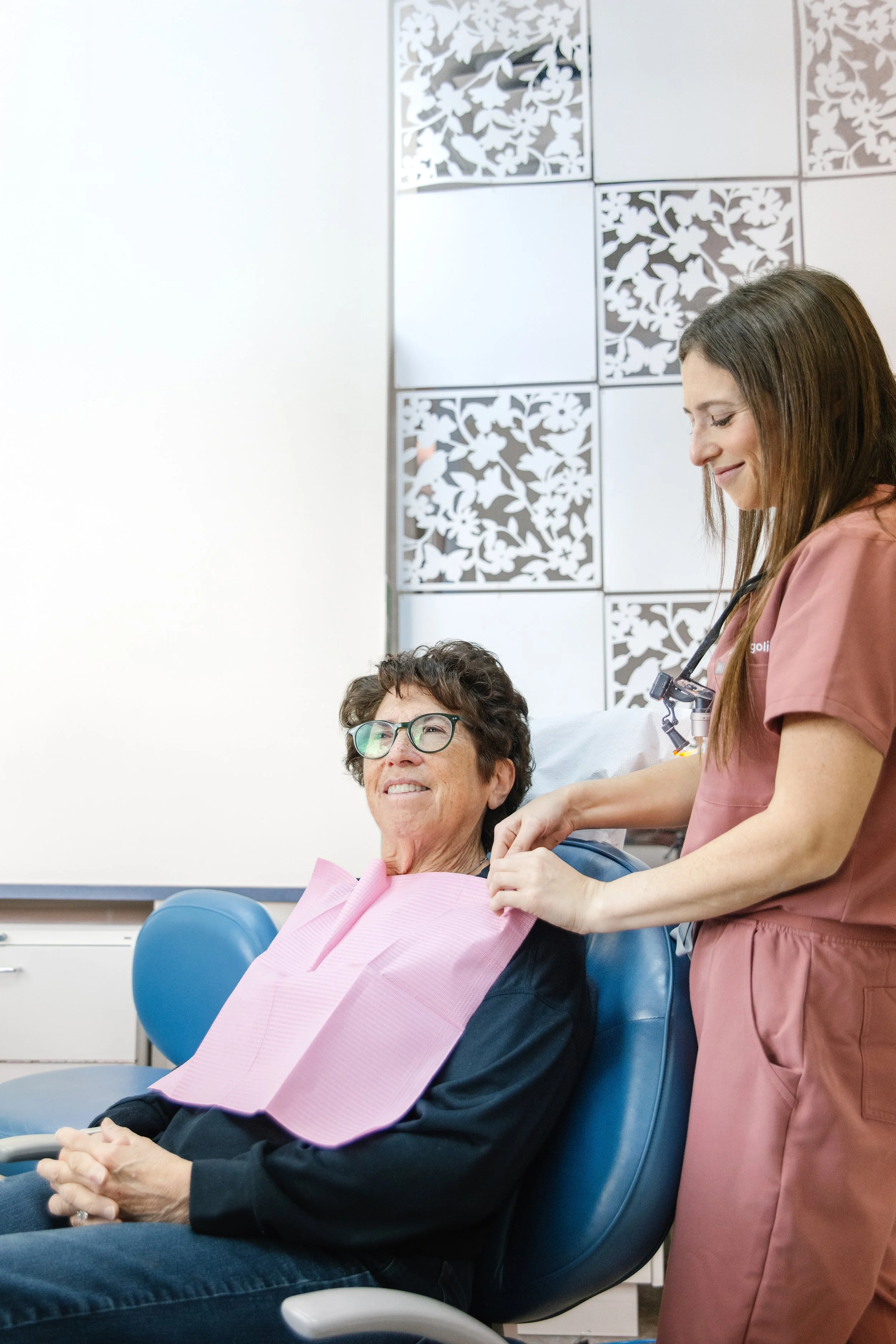 A woman in a pink medical gown sitting in a chair with a pink bib around her neck, smiling, while a nurse in pink scrubs stands beside her, preparing her for medical treatment in a clinical setting with decorative black and white wall panels.