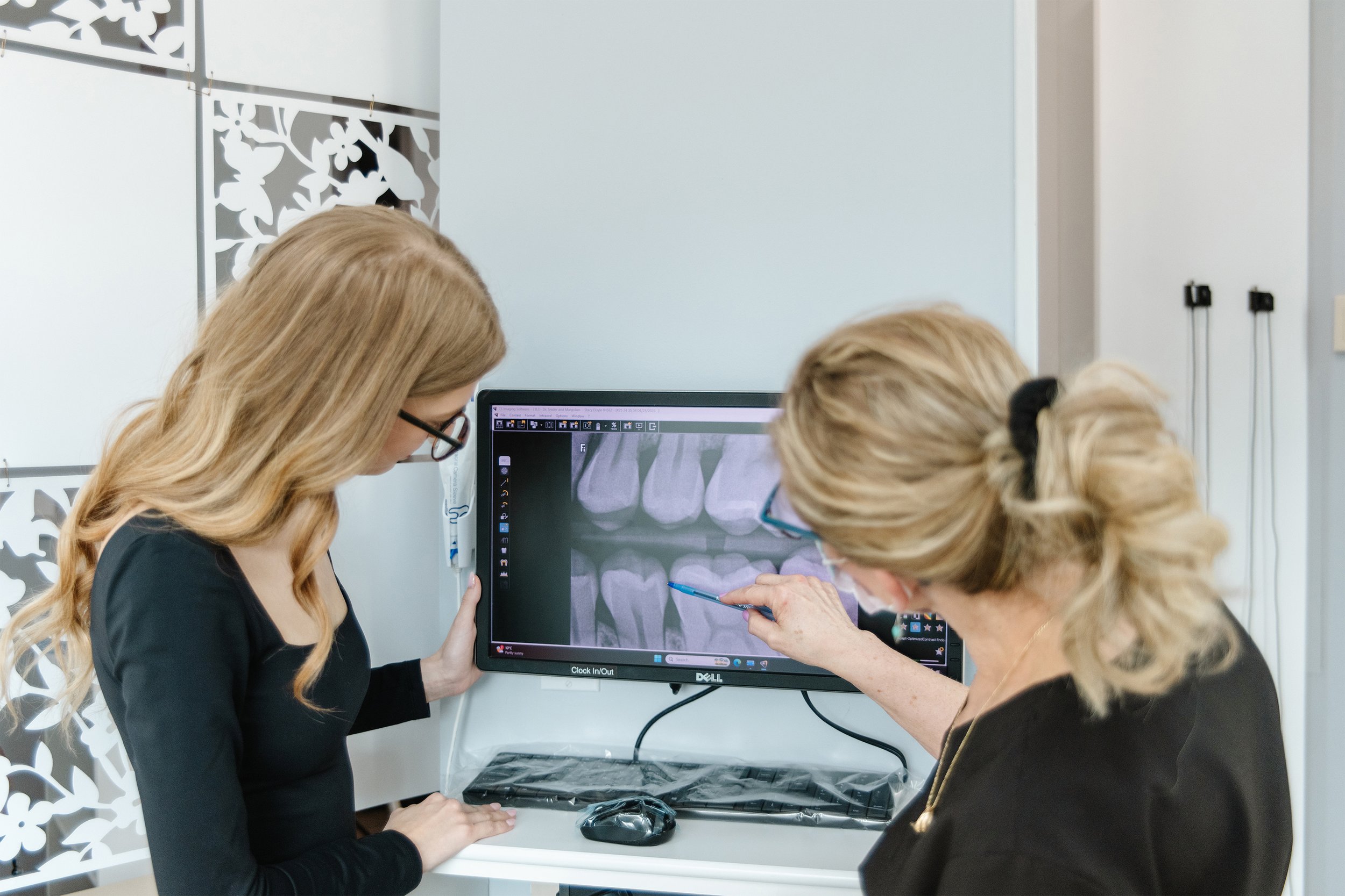 Two women, one younger and one older, are looking at dental X-ray images on a computer screen in a dental office. The older woman is pointing at the X-ray with a pen, and they are both wearing glasses.