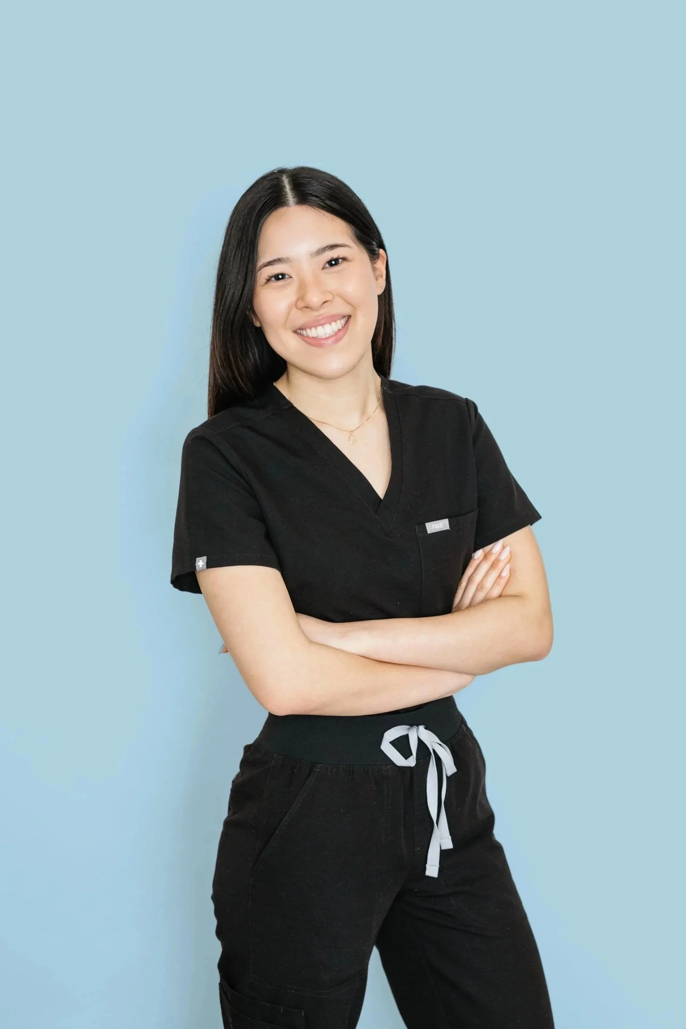 A young woman with long black hair, smiling, wearing a black scrub top and black drawstring pants, standing with arms crossed against a light blue background.
