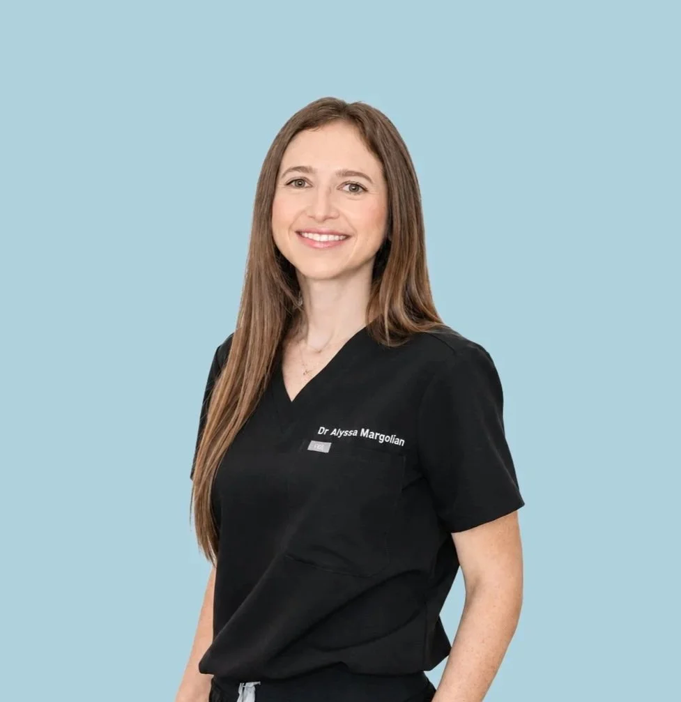 A woman with long brown hair wearing a black medical scrub top with the name "Dr Alyssa Margolian" embroidered on it, standing against a light blue background.