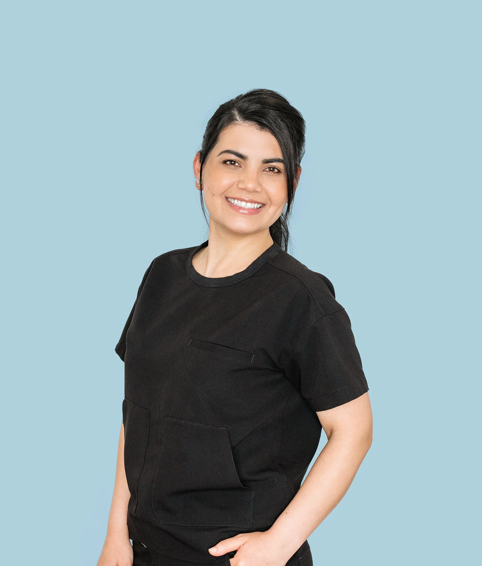 A woman with short dark hair, wearing a black scrub top, smiling against a light blue background.