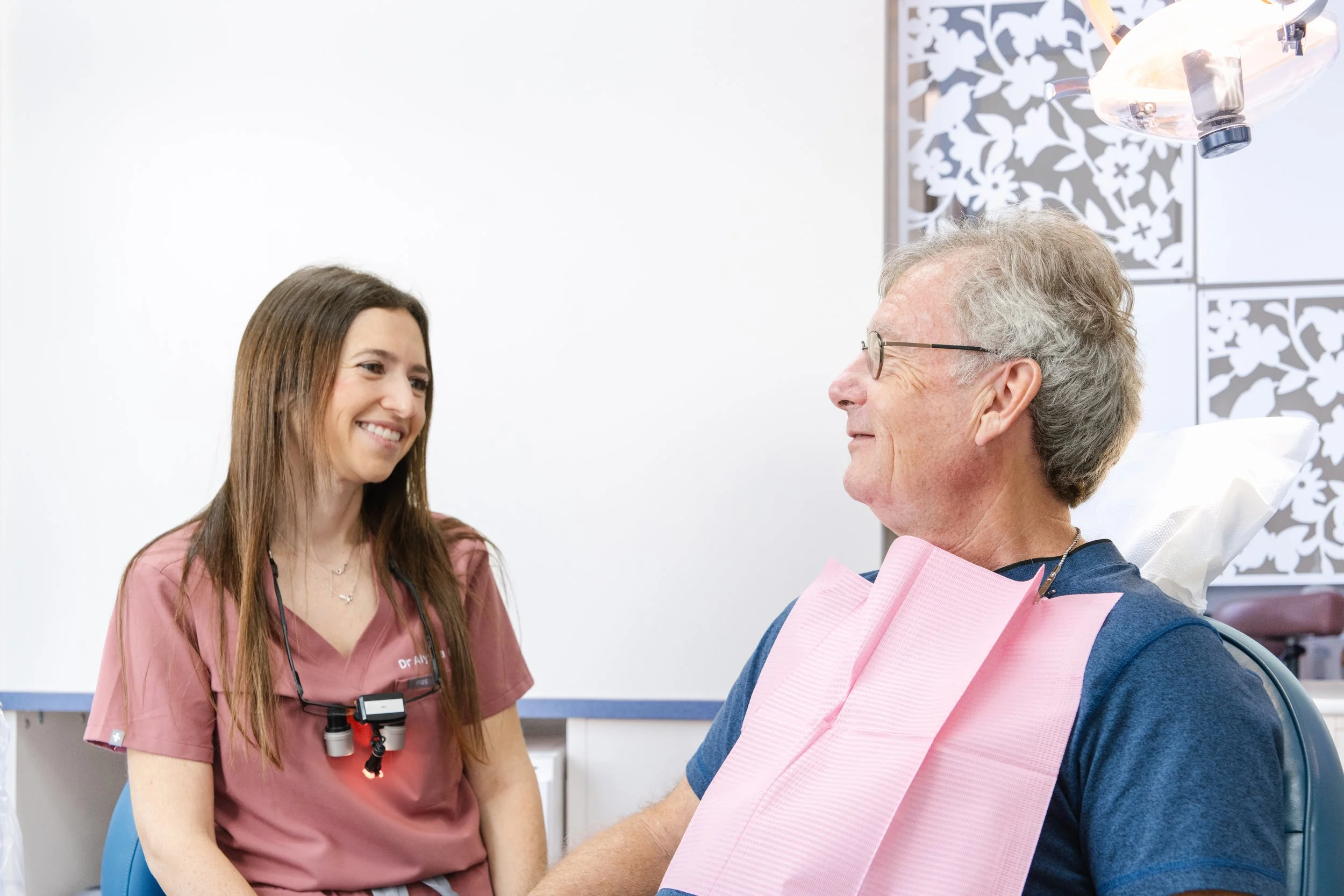 A young female healthcare worker in pink scrubs smiling and talking to an elderly man in a hospital bed with glasses, in a medical room with white walls and decorative panels in the background.
