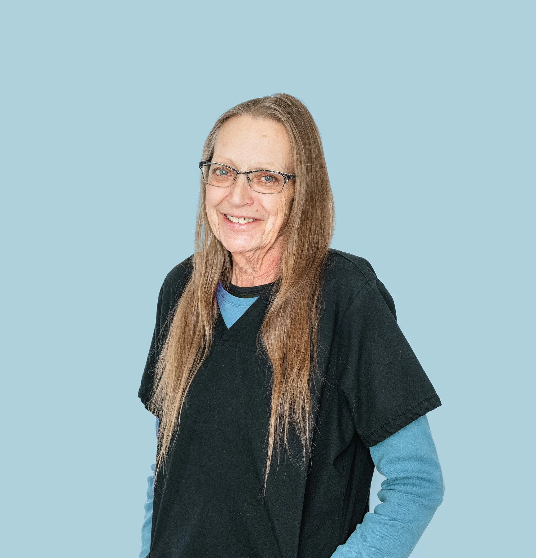 Portrait of a smiling woman with long brown hair, wearing glasses, a black top, and layered blue clothing, against a light blue background.