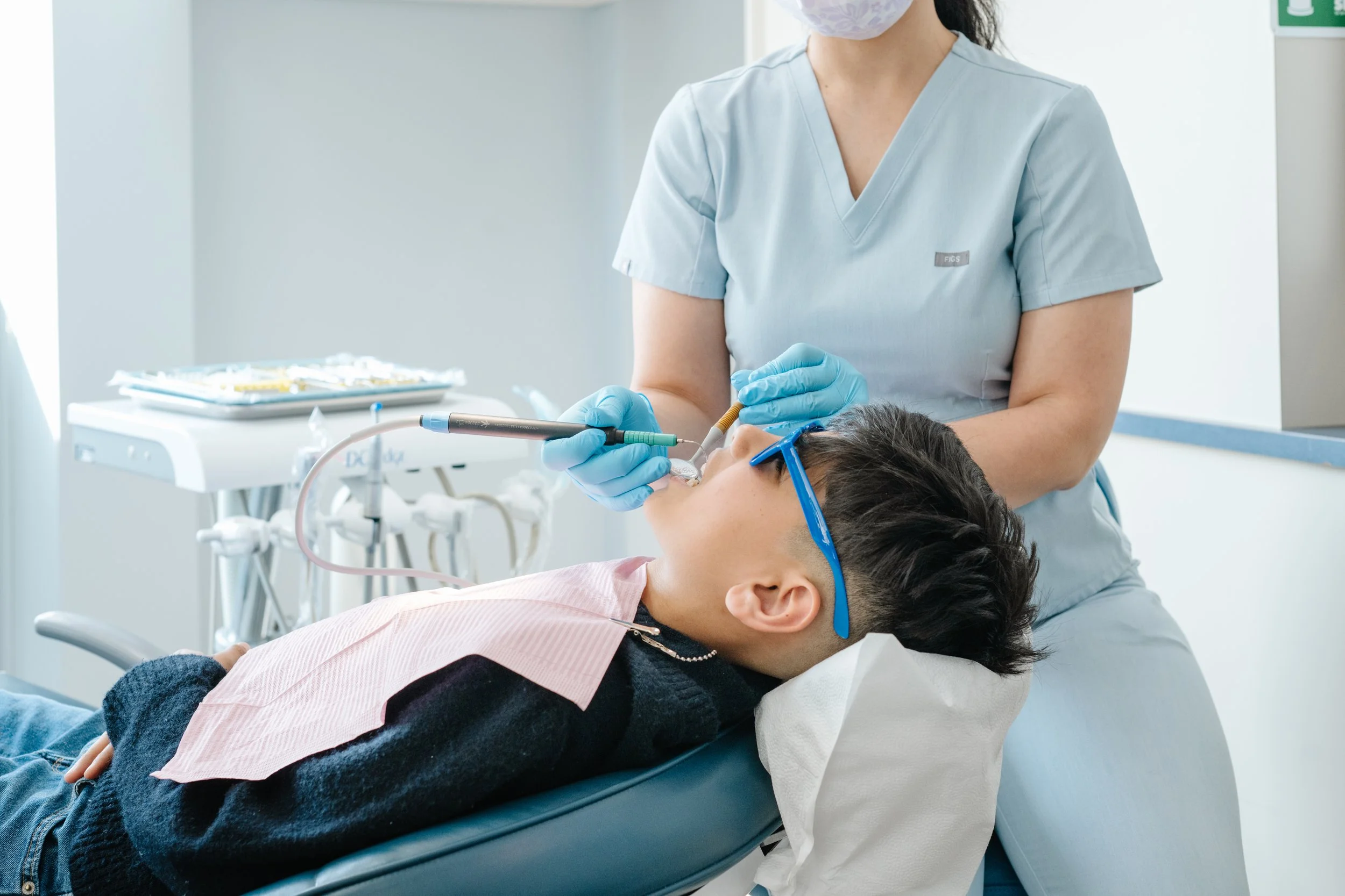 A healthcare worker in scrubs and a face mask performs dental work on a patient lying in a dental chair. The patient is wearing protective glasses and a bib, with dental tools and equipment visible in the background.