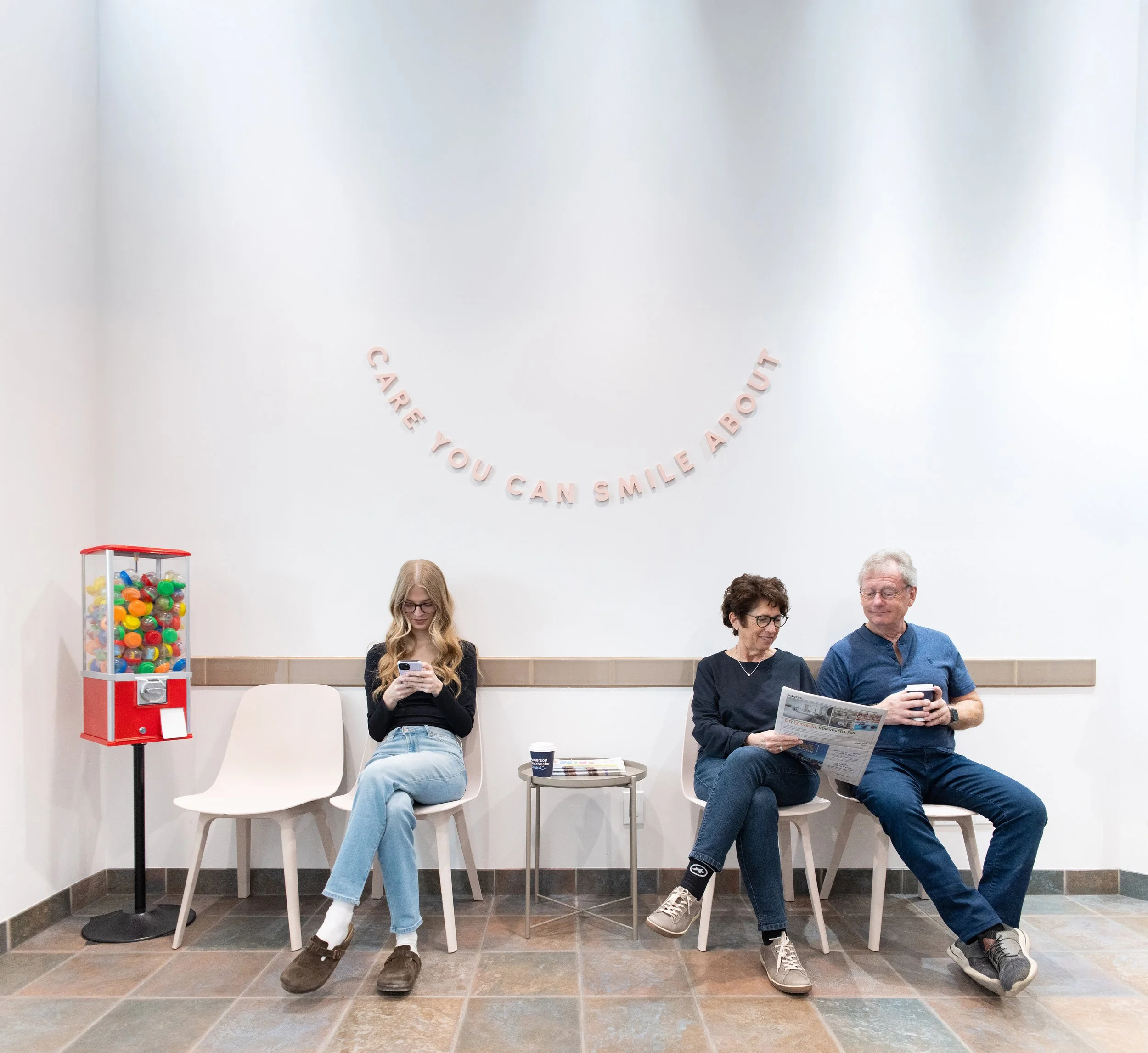Three people seated in chairs against a plain wall with pink lettering that says 'CARE YOU CAN SMILE ABOUT'. A vending machine filled with colorful gumballs is on the left. The first person is a young woman with long blonde hair, wearing glasses and a black top, looking at her phone. The second is an older woman with short dark hair, wearing glasses and a dark top, looking at the third person. The third is an older man with gray hair and glasses, wearing a blue shirt and jeans, reading a newspaper.