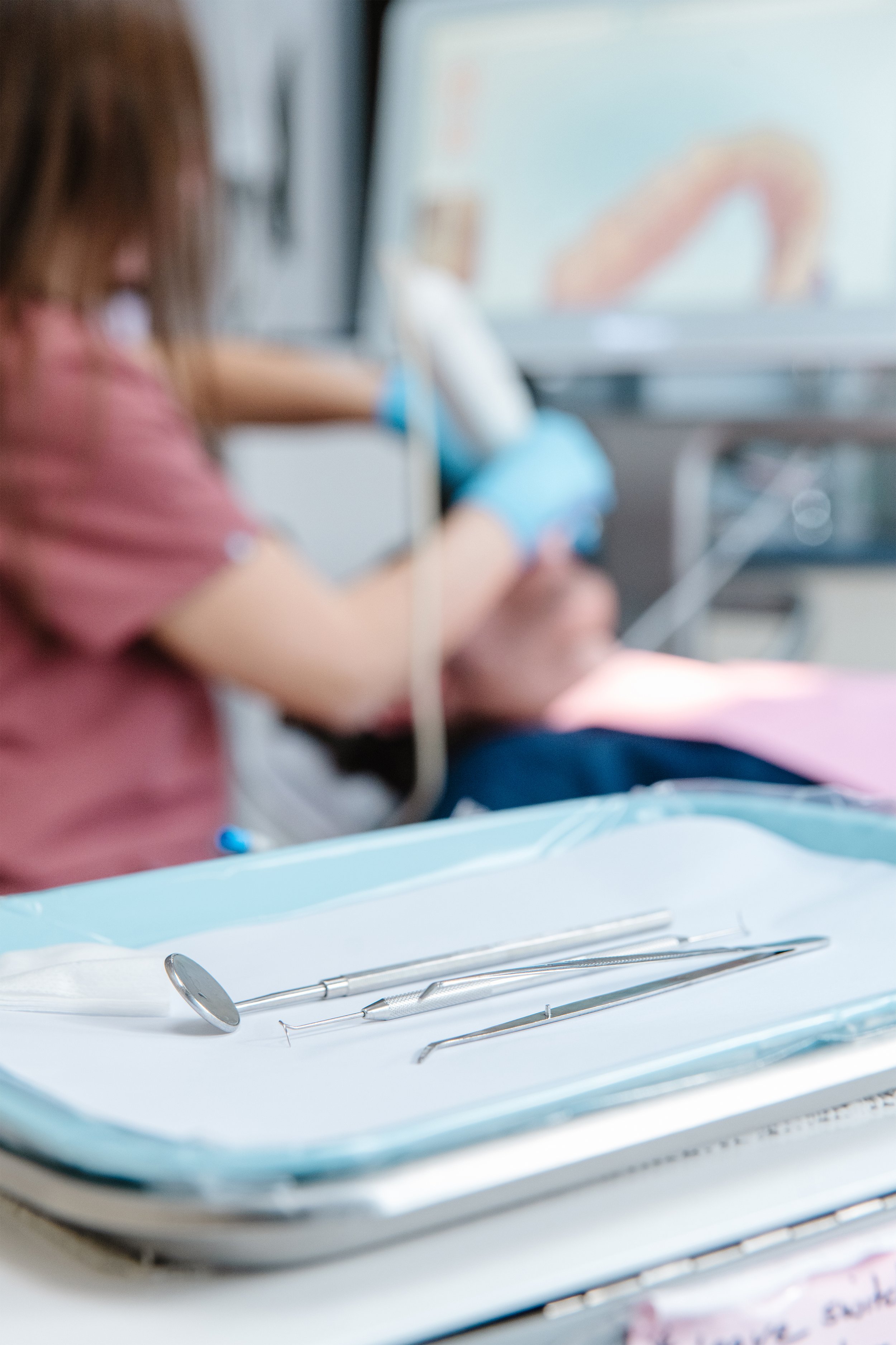 A dental tray with dental tools, including a mirror and explorer, placed on a white surface, with a blurred background of a person in a clinical setting.
