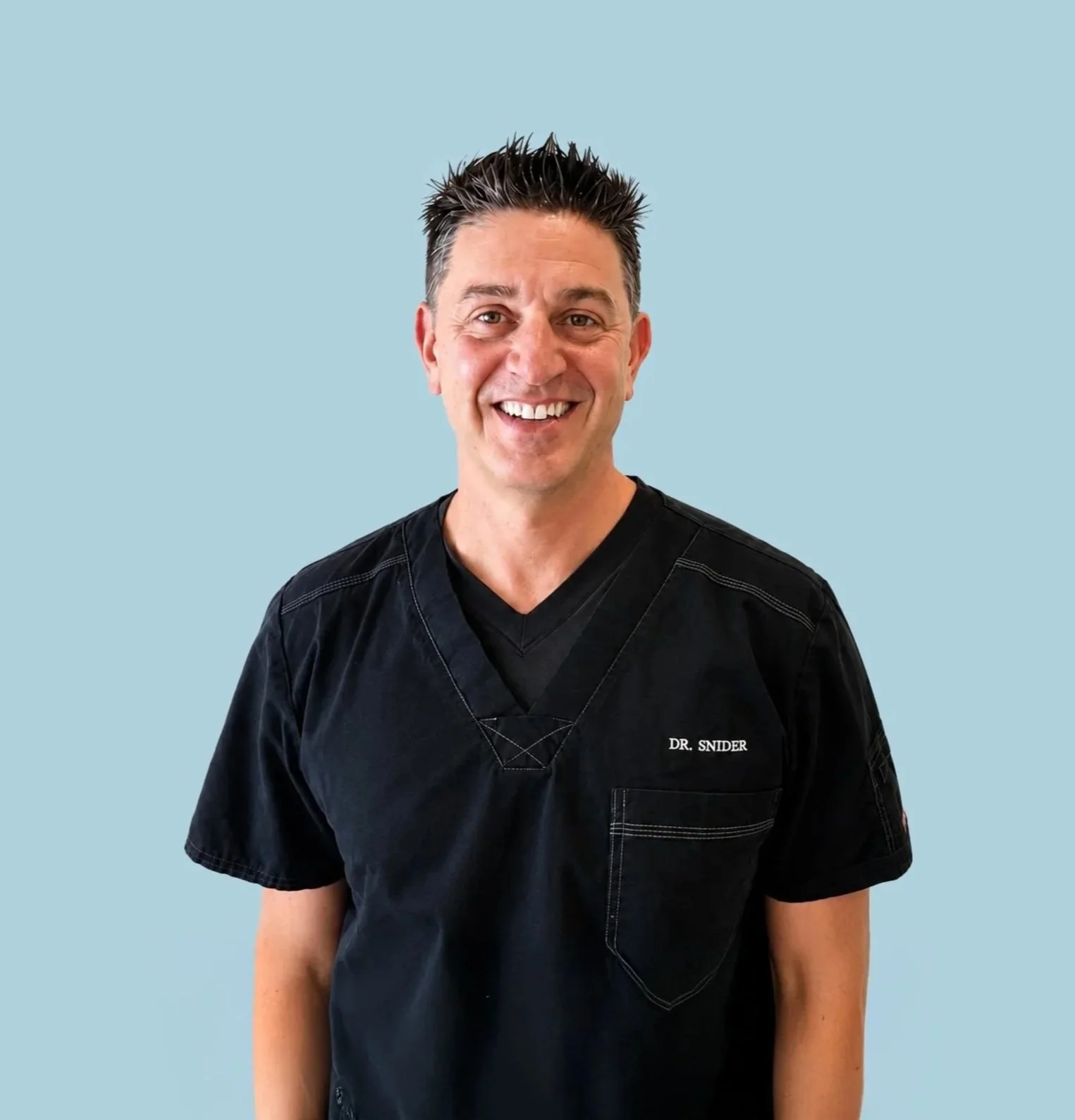 Portrait of a smiling male doctor wearing black scrubs with 'DR. SNIDER' embroidered on the chest, standing against a light blue background.