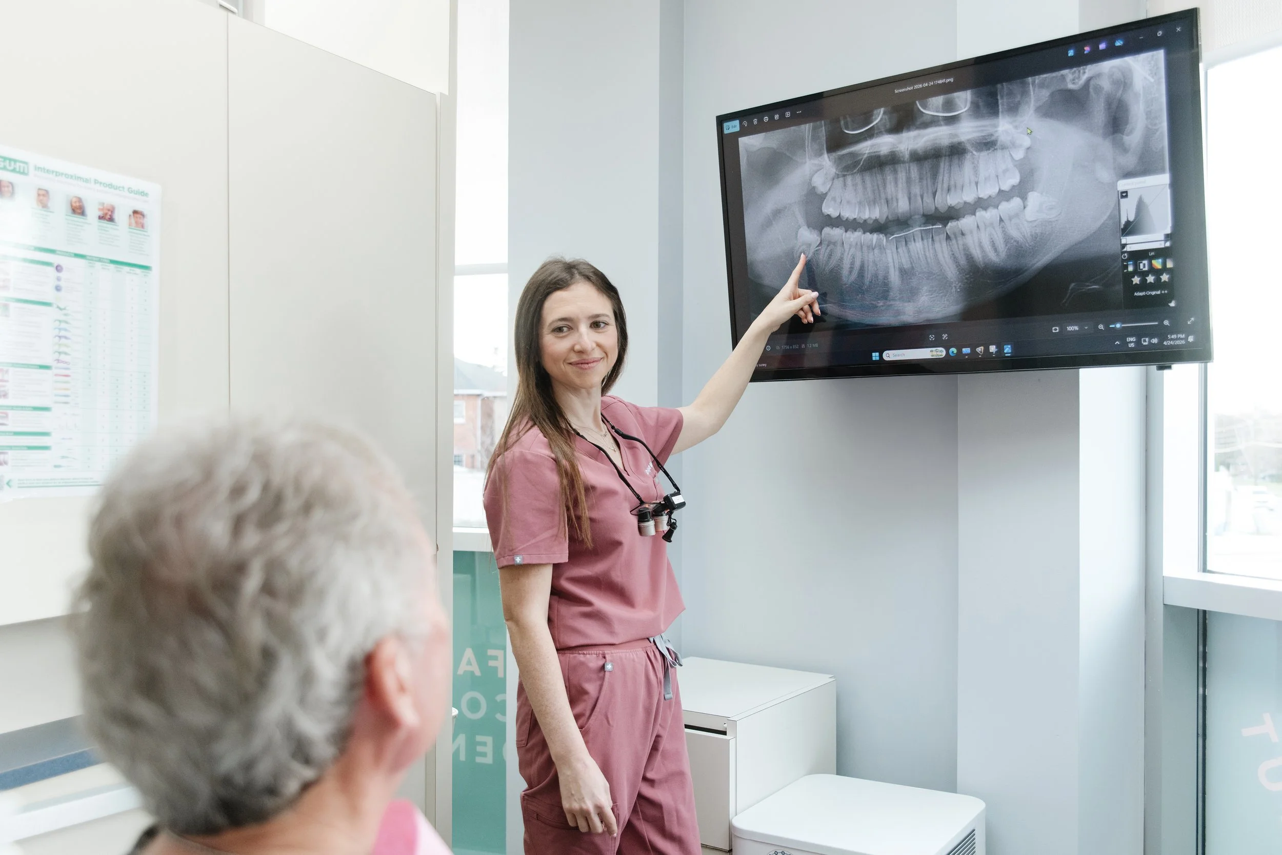 A female dentist in pink scrubs points at a dental X-ray displayed on a monitor, while an elderly patient with gray hair looks on in a dental office.