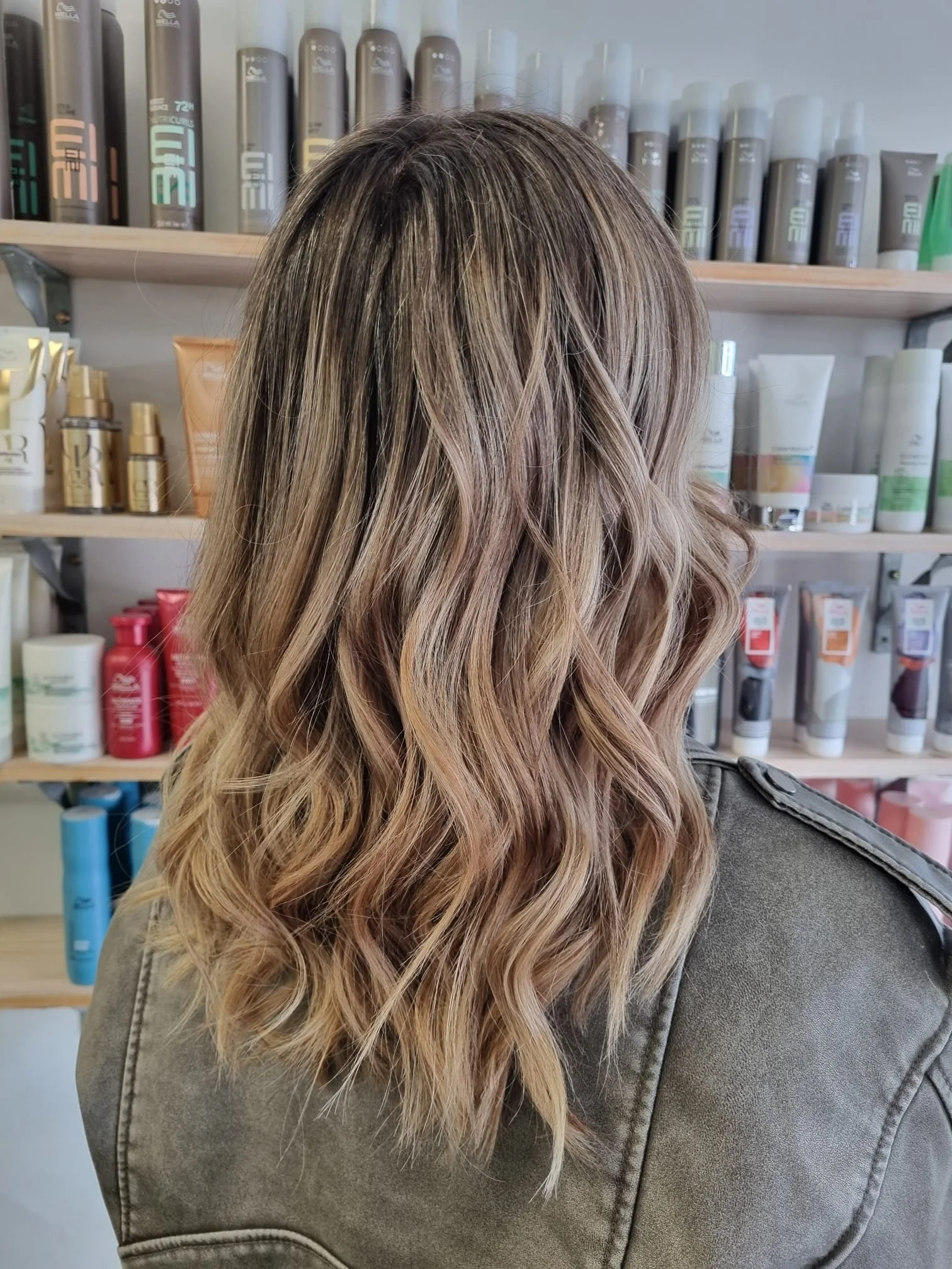 Woman with shoulder-length wavy hair in shades of blonde and light brown, viewed from the back in a salon or store with shelves of hair and beauty products.